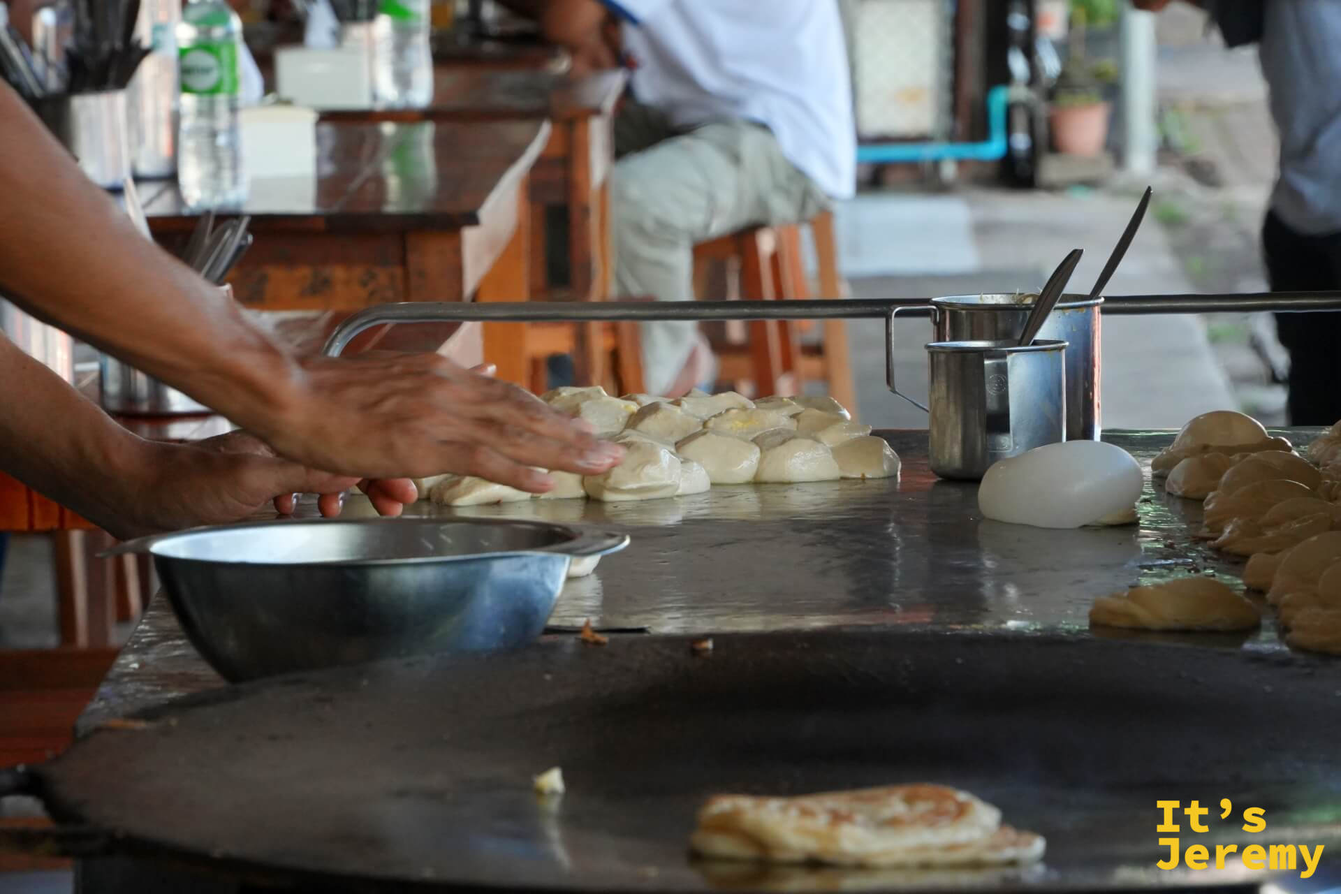 雙手將麵糰鋪展在鐵板上，右側的圓鐵盤上已有一張 Roti 開始煎制