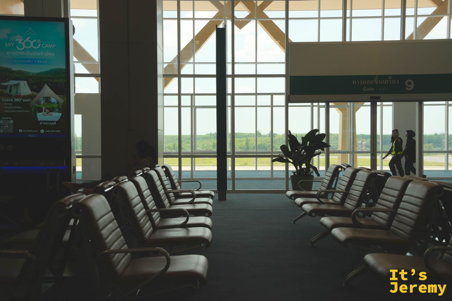喀比機場候機室,Gate 9 登機門,大片玻璃窗正對停機坪與遠方綠意,兩名地勤人員走過