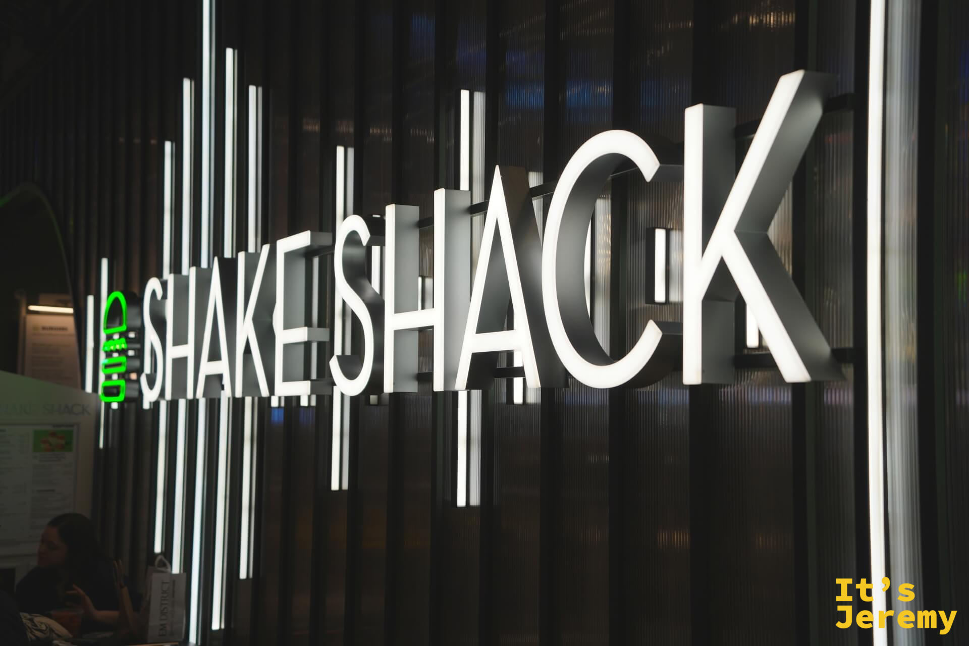 Shake Shack 發光招牌特寫,白色大字在黑色縱條格柵牆上,左側綠色漢堡 logo