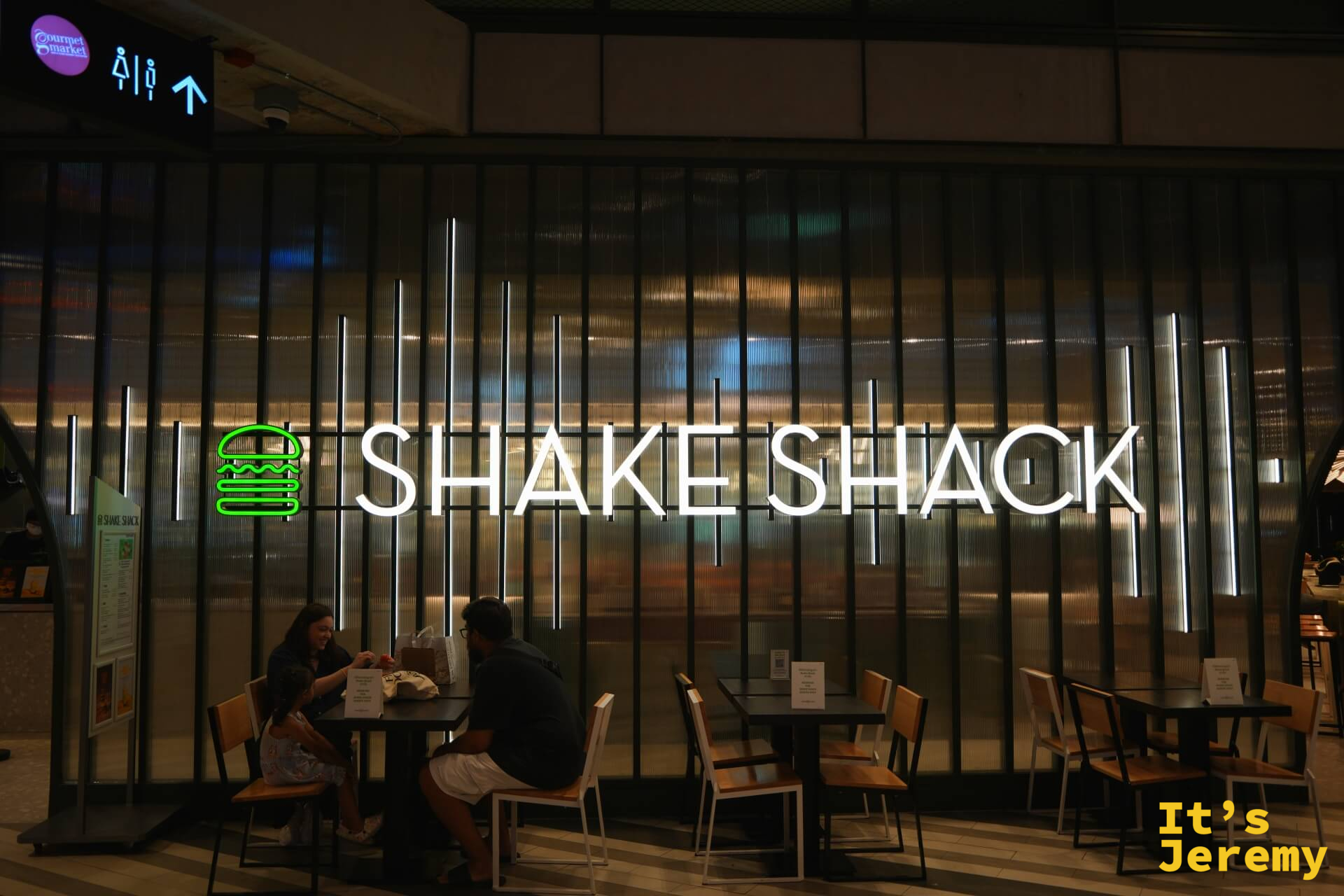 Shake Shack Emsphere 店面全貌,招牌打亮,顧客坐在店外木椅桌用餐,上方 Gourmet Market 指示牌