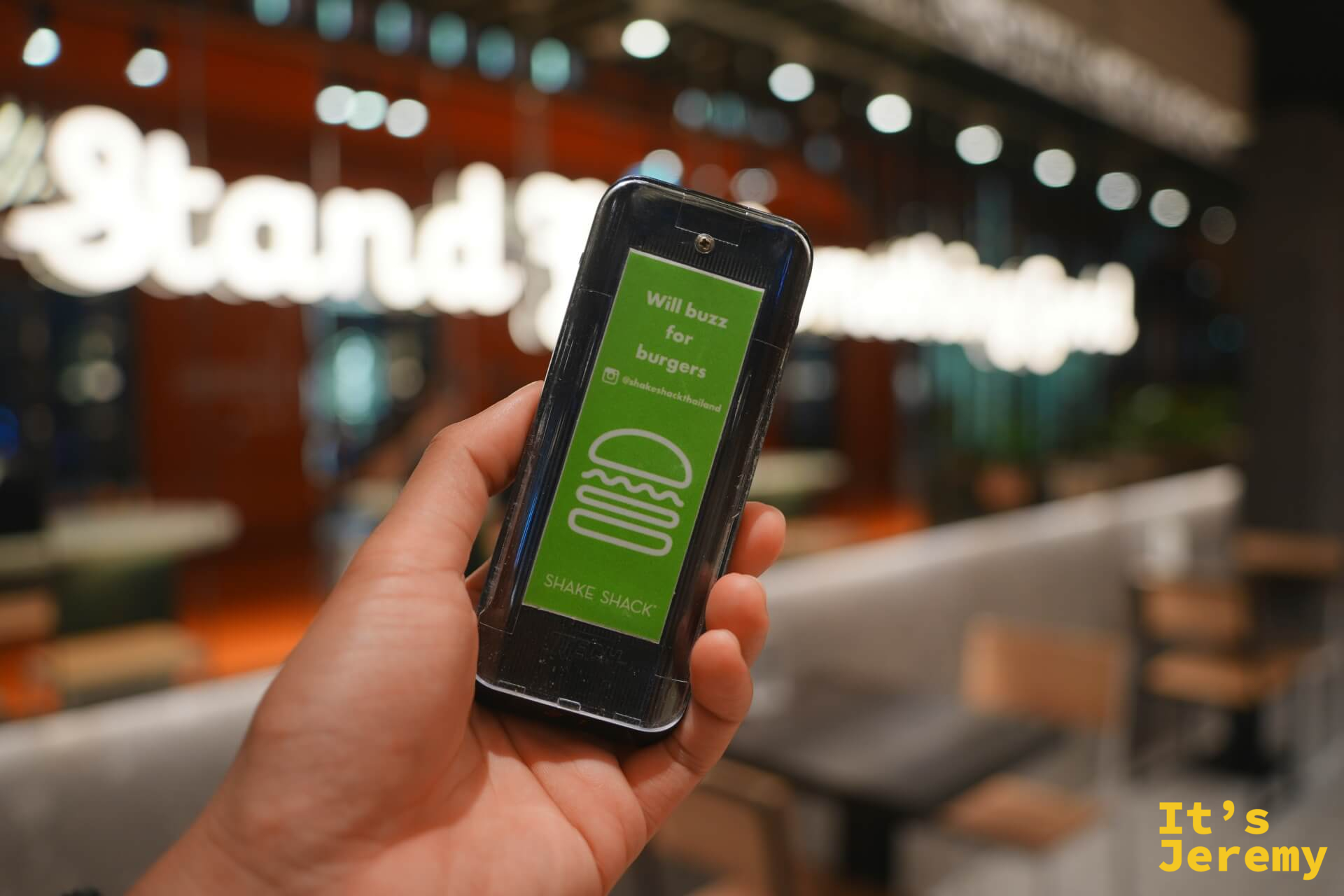 手持 Shake Shack 取餐震動器,正面印「Will buzz for burgers」和漢堡圖案,背景是店內空間