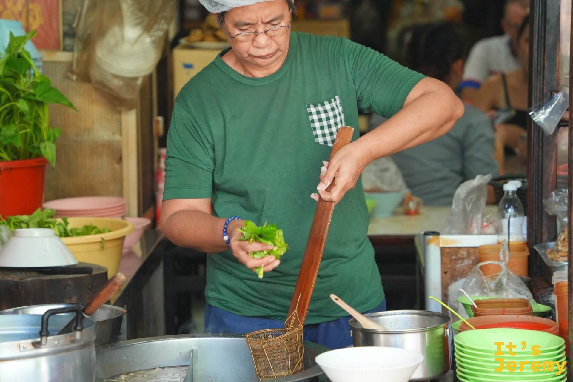 廚師手持長木柄鍋勺,在鍋前燙菜備料,背景是大型湯鍋與整排碗碟