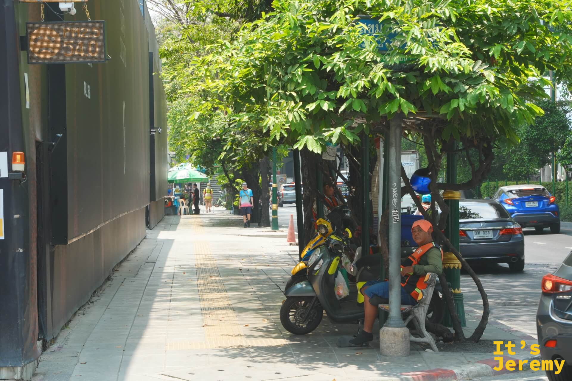 路旁建築牆上的另一塊 PM2.5 電子看板顯示數值 54.0,下方可見幾個工人在僅有的樹蔭下納涼