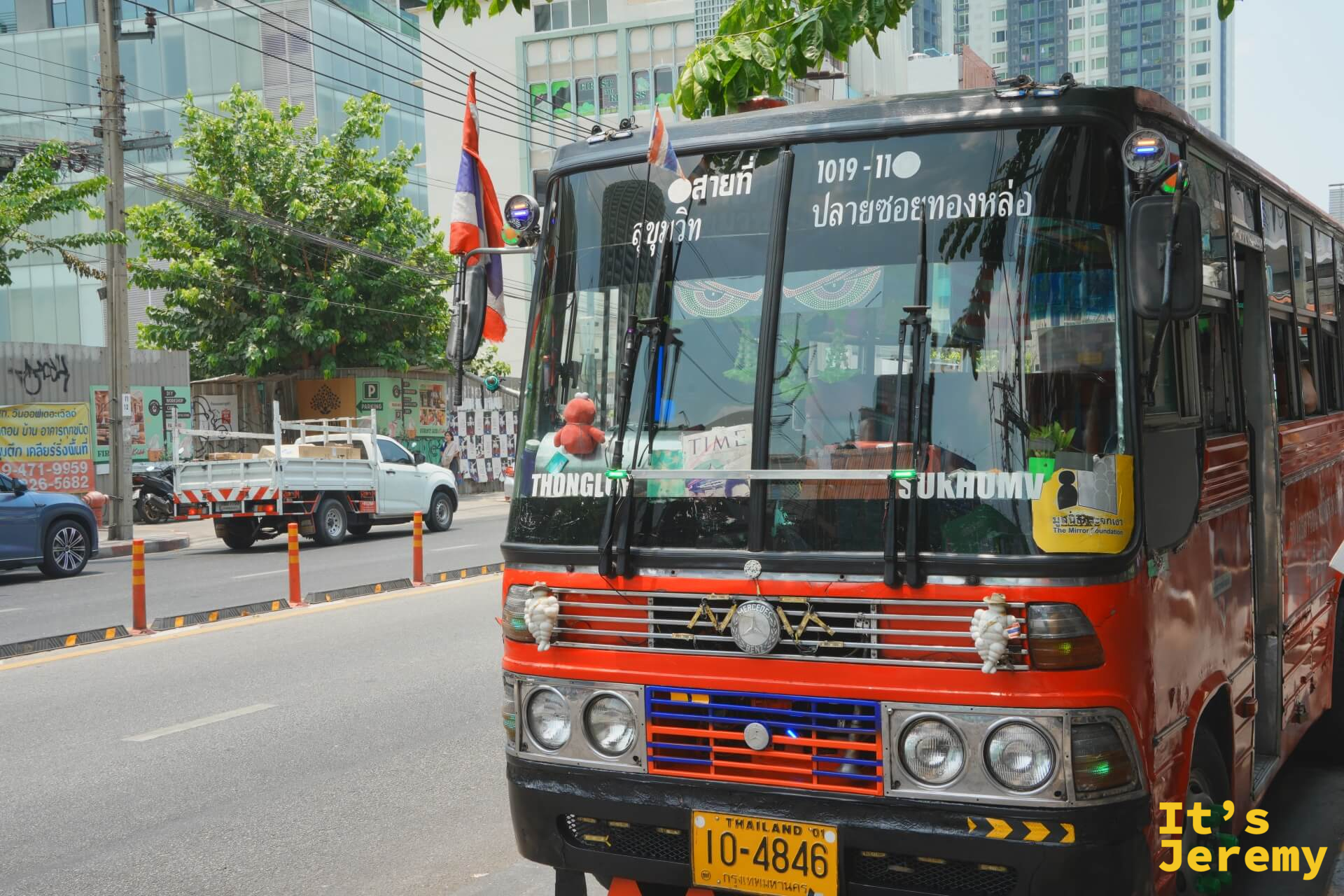 一輛紅黑色的曼谷老公車停在路旁,車身已有些年紀,和不遠處的 BTS 高架橋形成有趣的時代反差