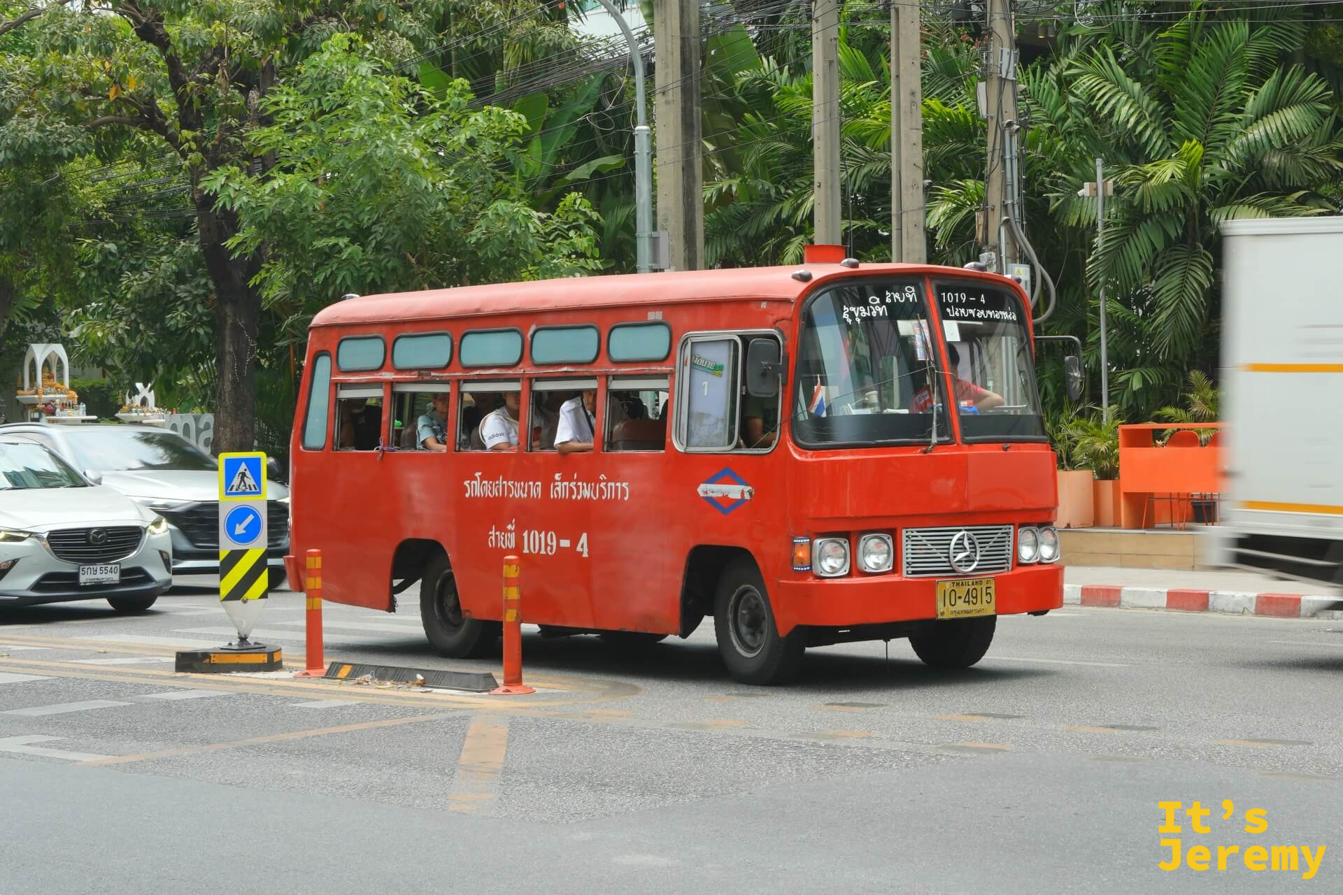 一輛小型紅色曼谷公車穿過路口,舊款車身的顏色在陽光裡顯得有點褪,走進熱氣中