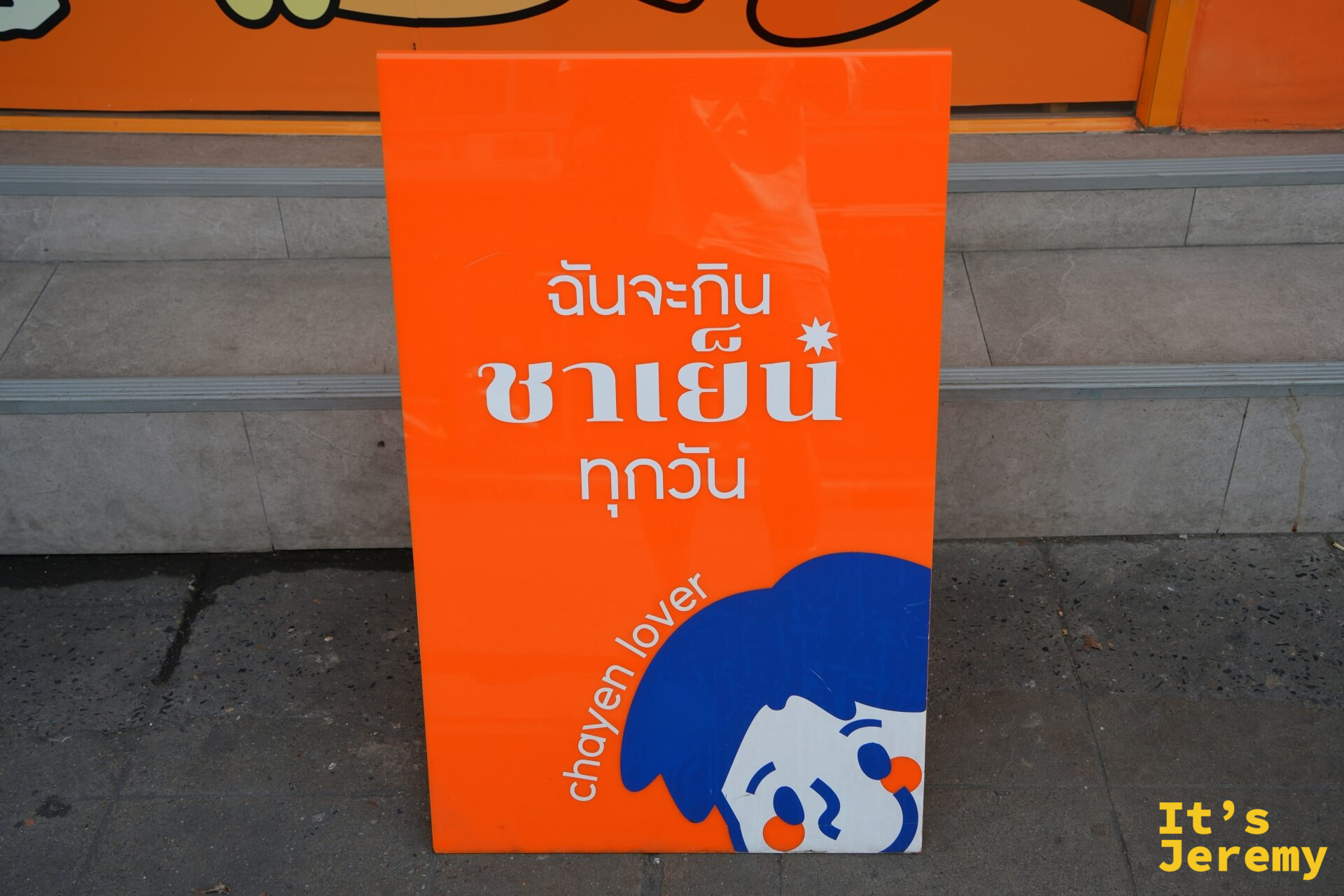 橘底白字的 A 字板,寫著「ฉันจะกิน ชาเย็น ทุกวัน」,chayen lover 角色從下方探出頭來