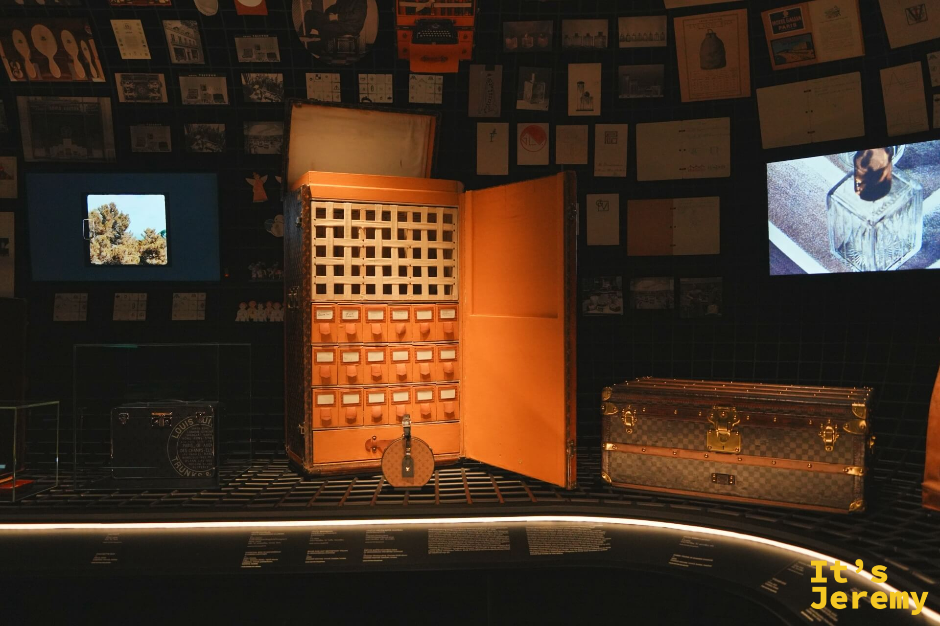 橘色大型行李箱展開，內部設計成多層抽屜收納格，旁邊陳列棋盤格 LV 行李箱，牆面貼滿歷史廣告
