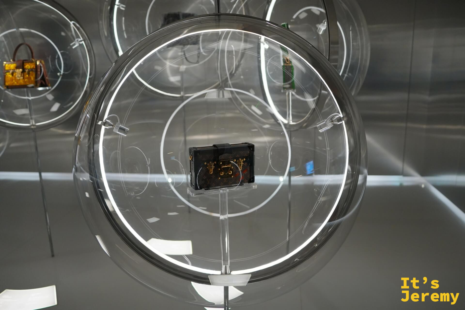 圓形壓克力展示泡泡，中央固定著黑色 LV Petite Malle 迷你行李箱，背景多個類似展示球延伸陳列