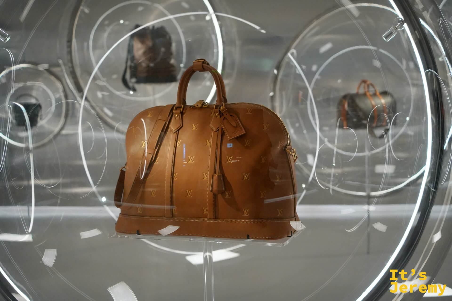圓形壓克力展示泡泡，中央是棕色 monogram 皮革 Keepall 旅行包，聚光燈透過球面折射出柔和光暈