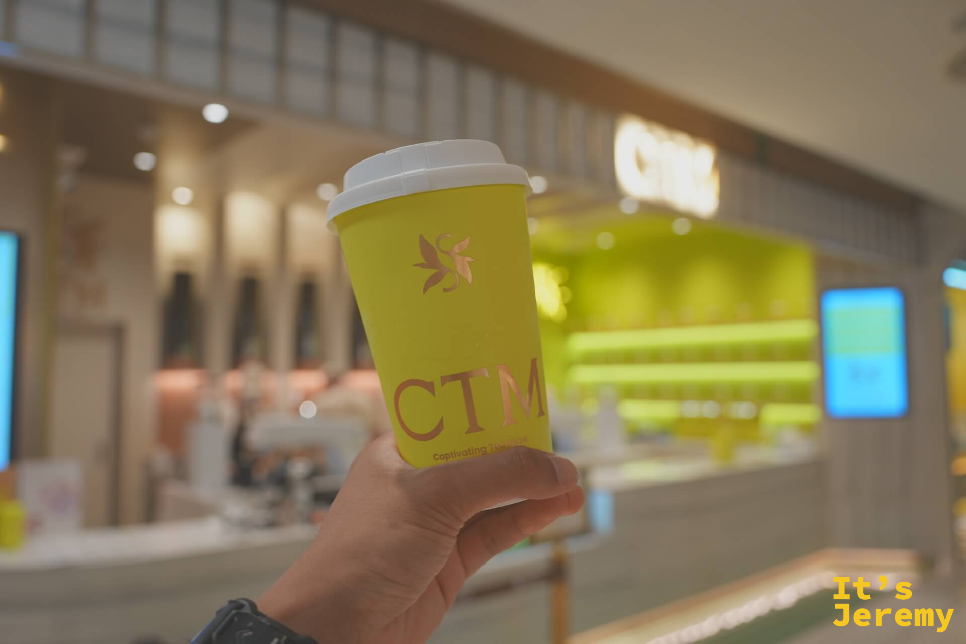 手持 CTM 招牌黃色茶飲杯,顏色飽和度很高,在 Central Park Bangkok 的空間背景裡顯得很突出