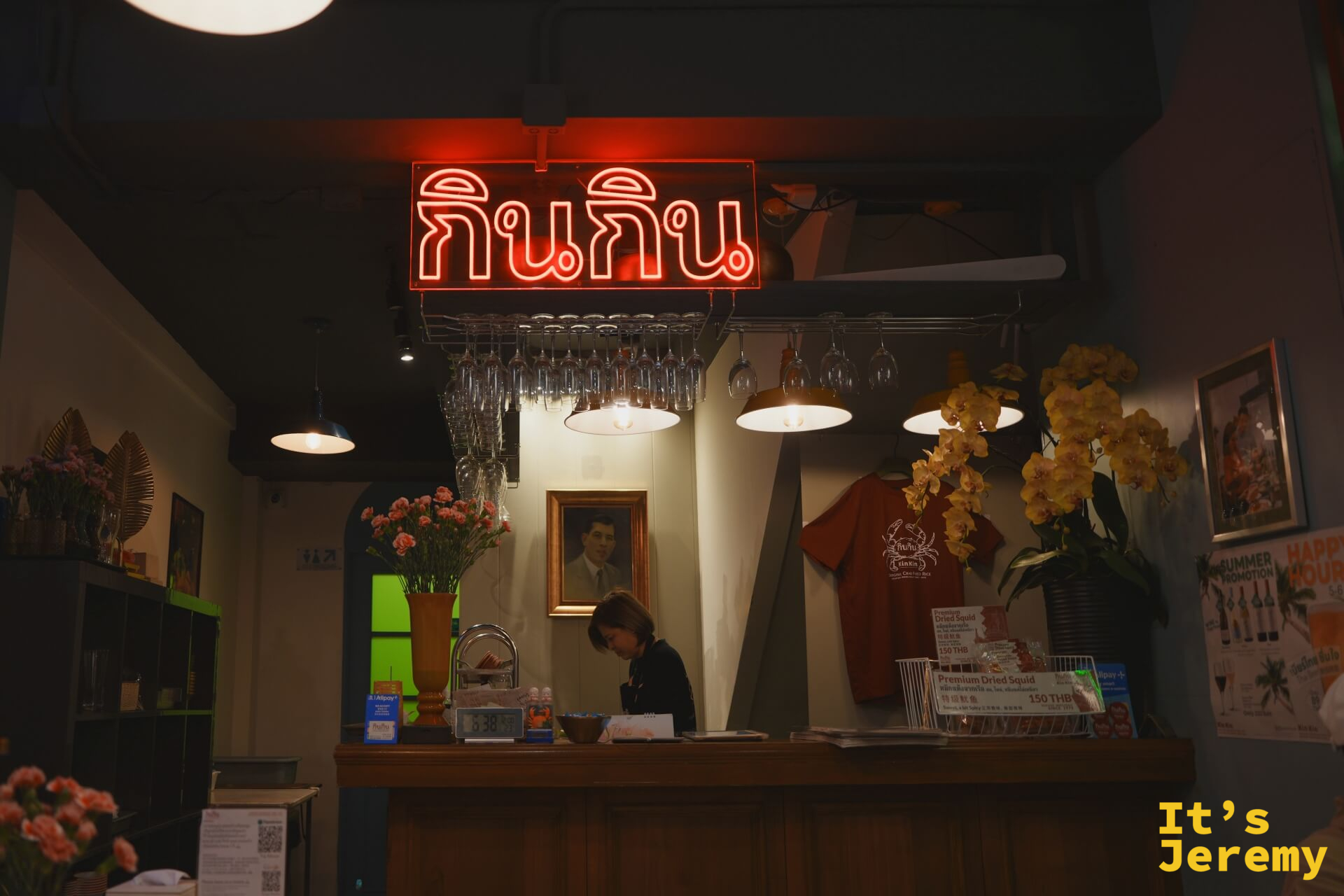 吧台上方的紅色霓虹「กินกิน」字樣，後方掛著一幅老照片，收銀台旁展示著自家品牌周邊