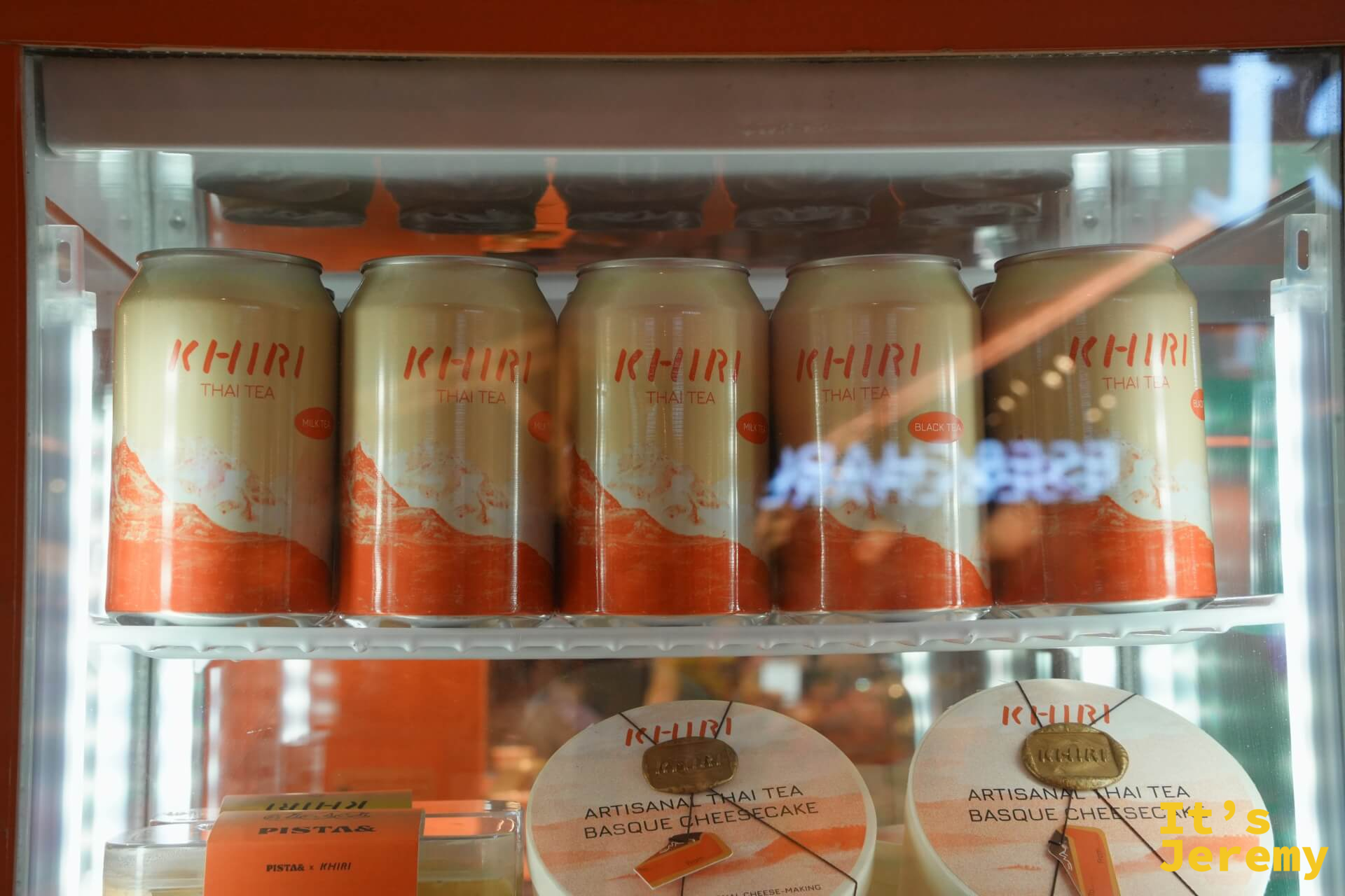 展示冰箱近景：橘色 KHIRI 罐裝（Milk Tea 與 Black Tea 款）與 Artisanal Thai Tea Basque Cheesecake