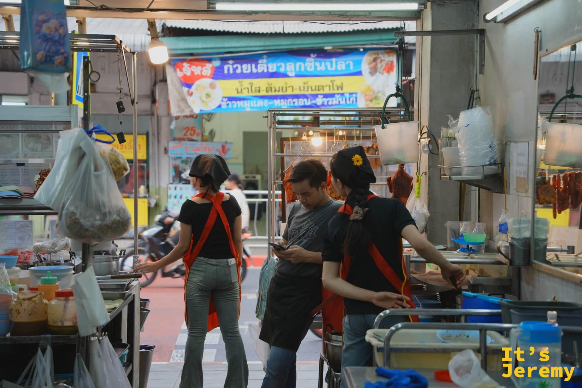 店內景象:穿黑衣紅圍裙的服務員和客人在點餐,上方是泰文招牌「เจ้าหลงก๊วยเตี๋ยวลูกชิ้นปลา」
