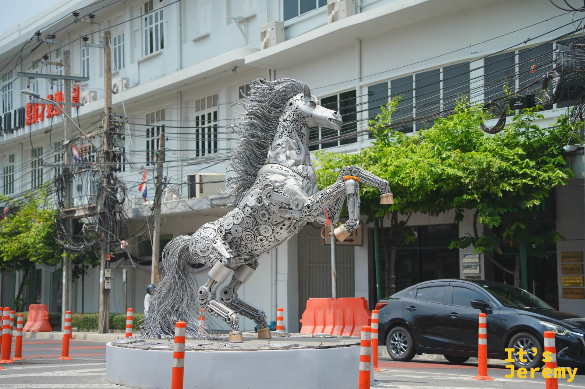 路口擺放的大型廢鐵件拼組金屬馬雕塑,揚蹄姿態,後方是白色建築