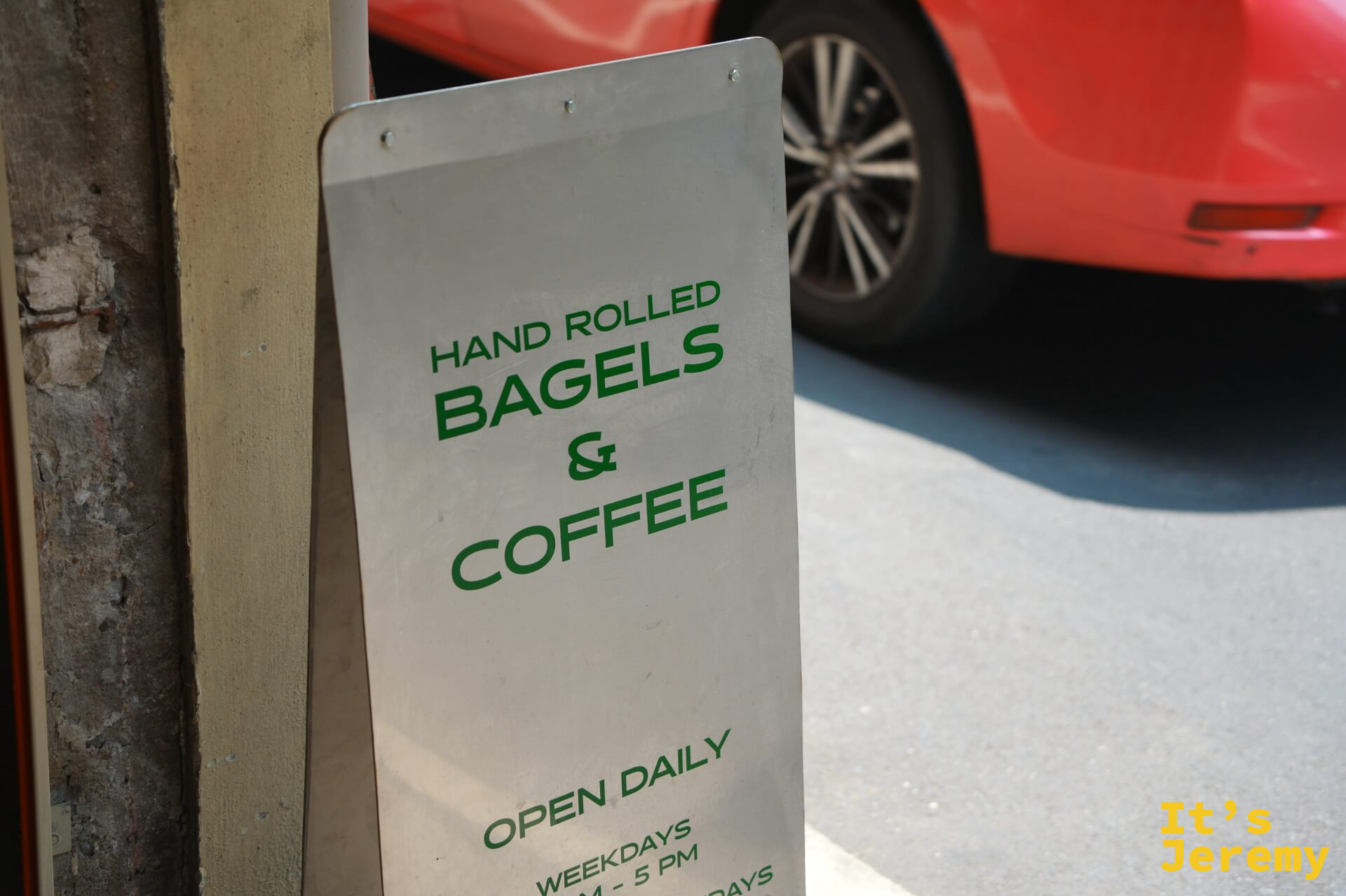 店門口的金屬立牌：「HAND ROLLED BAGELS & COFFEE, OPEN DAILY」，背景是路邊停放的紅色汽車