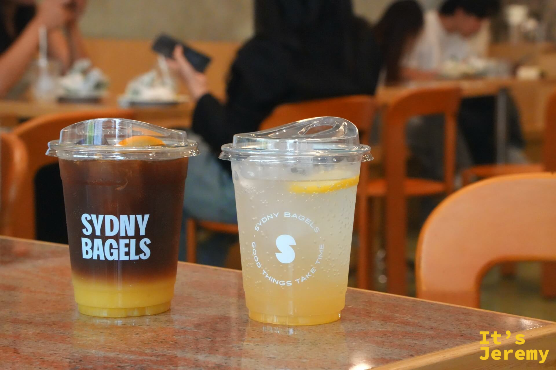 桌面上的兩杯飲料：「SYDNY BAGELS」字樣的黑咖啡與印有圓形 logo 的氣泡飲並排