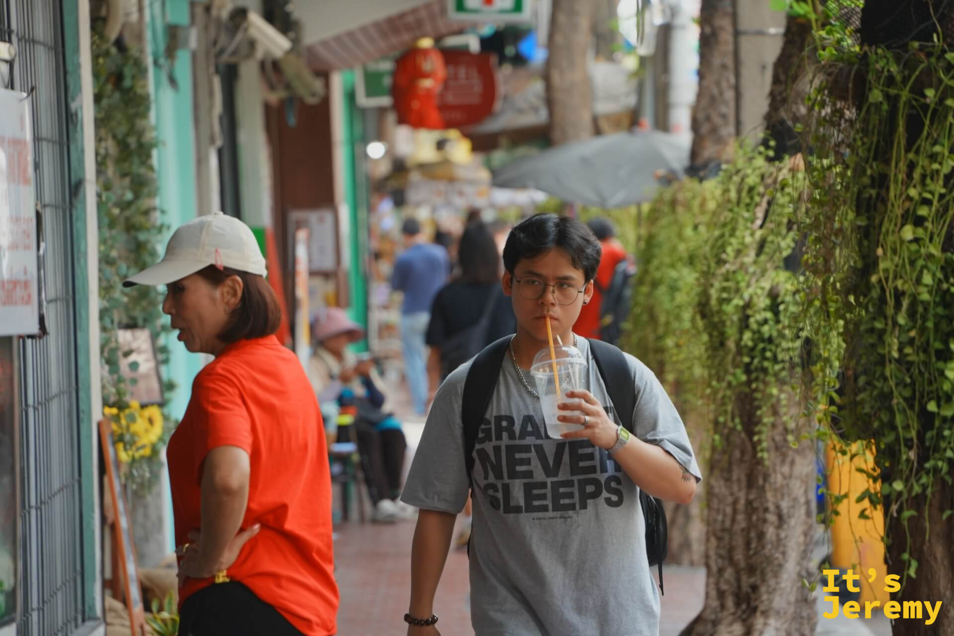 年輕男子邊走邊用吸管喝飲料,T 恤印著「Grains Never Sleeps」,背後是懸掛植物的廊道