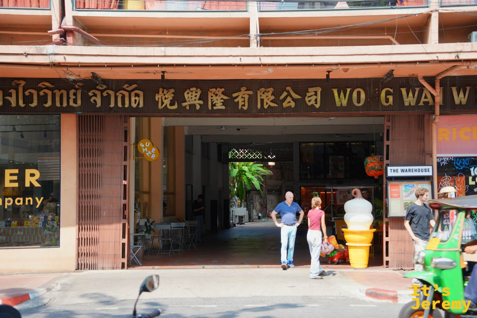THE WAREHOUSE Bangkok 入口外觀:老倉庫門洞,旁邊掛著「悅興隆有限公司」招牌,THE WAREHOUSE 招牌在斜後方,一台嘟嘟車停在外
