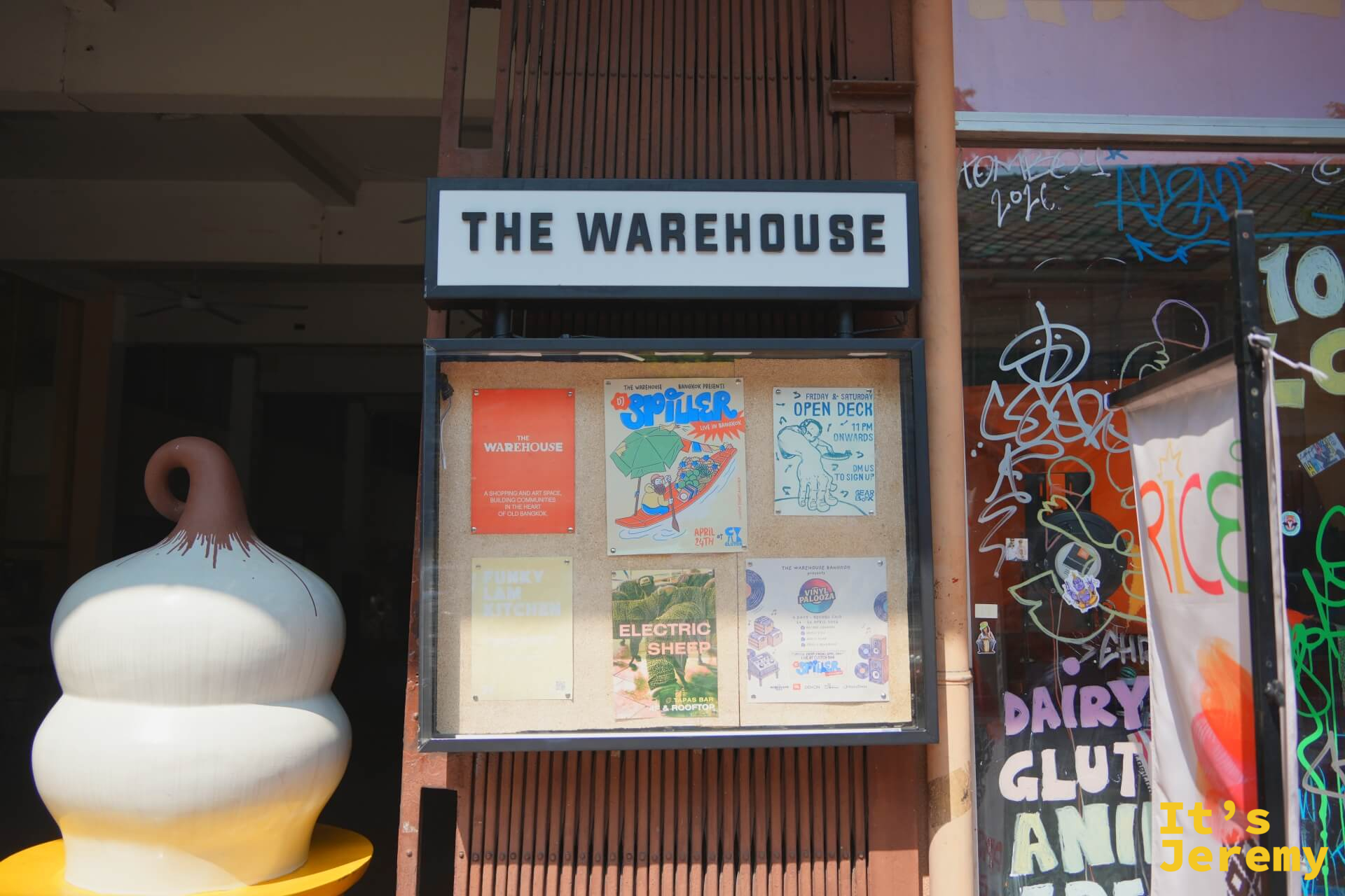 THE WAREHOUSE 正面外觀:「THE WAREHOUSE」白字招牌,下方是活動佈告欄,旁邊有白色陶瓷裝置藝術