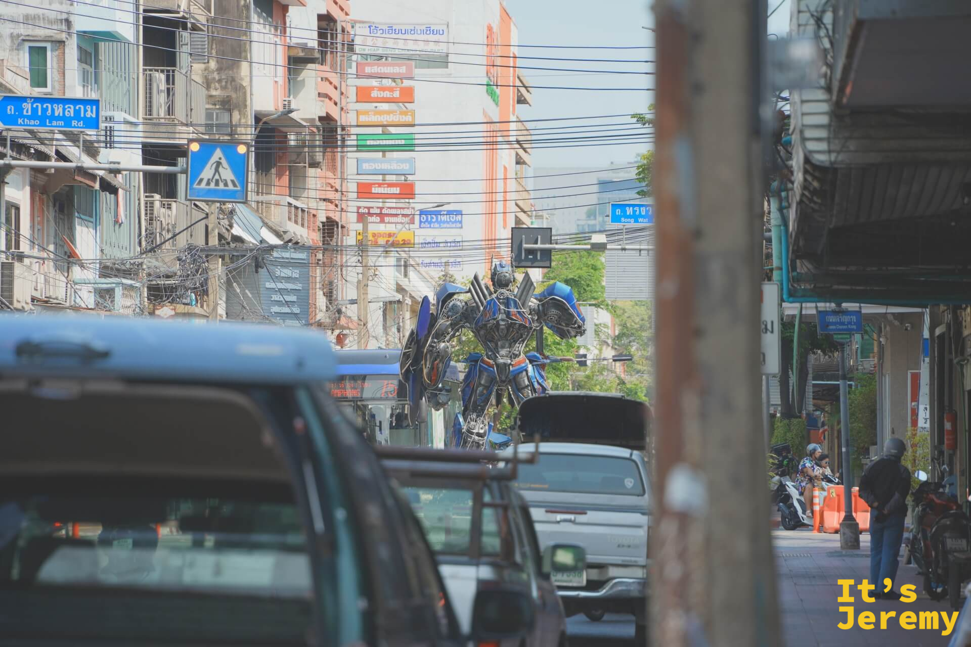 從遠處透過車流看到嵩越路路口,擎天柱雕塑在車輛之間矗立,路牌標示「Khao Lam Rd.」和「Song Wat」