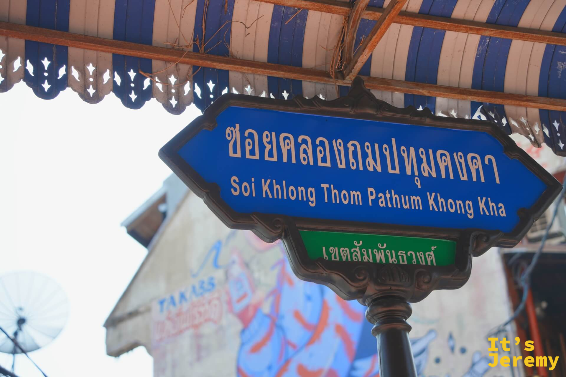藍色泰英雙語路標「Soi Khlong Thom Pathum Khong Kha」,背後建築牆面是密集的塗鴉字體
