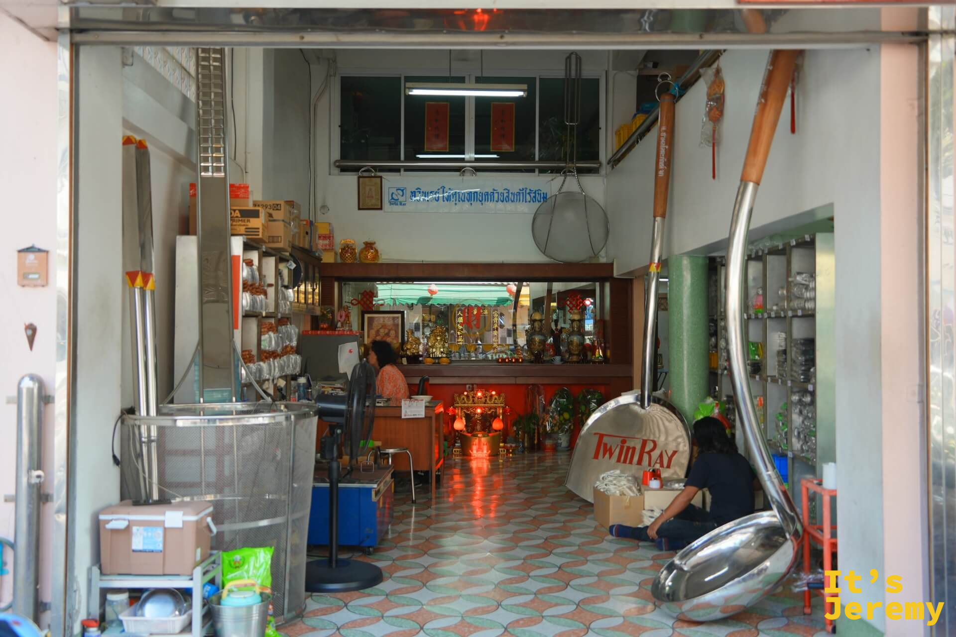廚具行入口:超大型不鏽鋼炒鏟和舀勺靠在門邊,店內可見神桌、磁磚地板和女店員