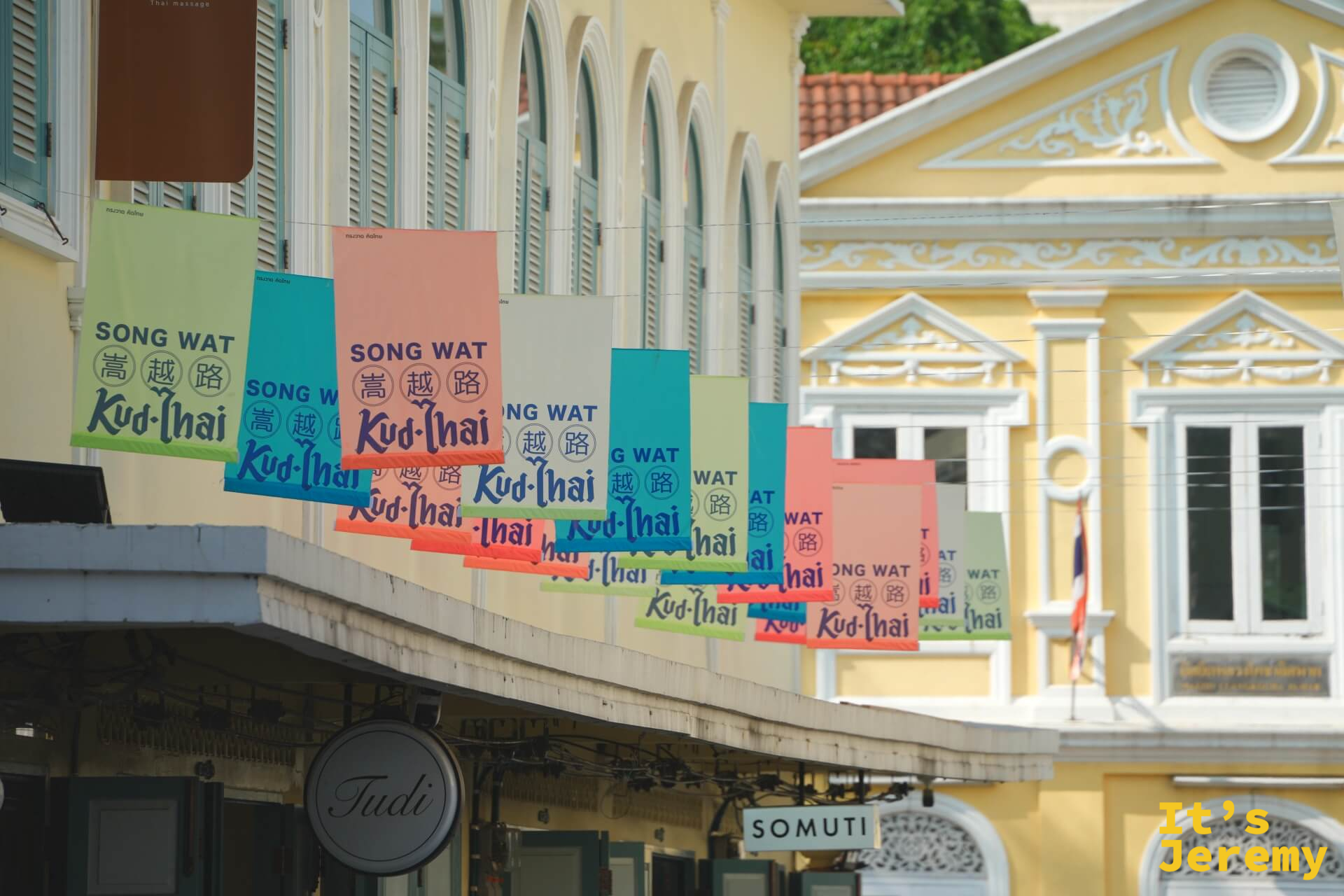 多色「SONG WAT 嵩越路 Kud-Thai」旗幟密排在黃色新古典主義建築前,「Judi」與「SOMUTI」招牌清晰可見