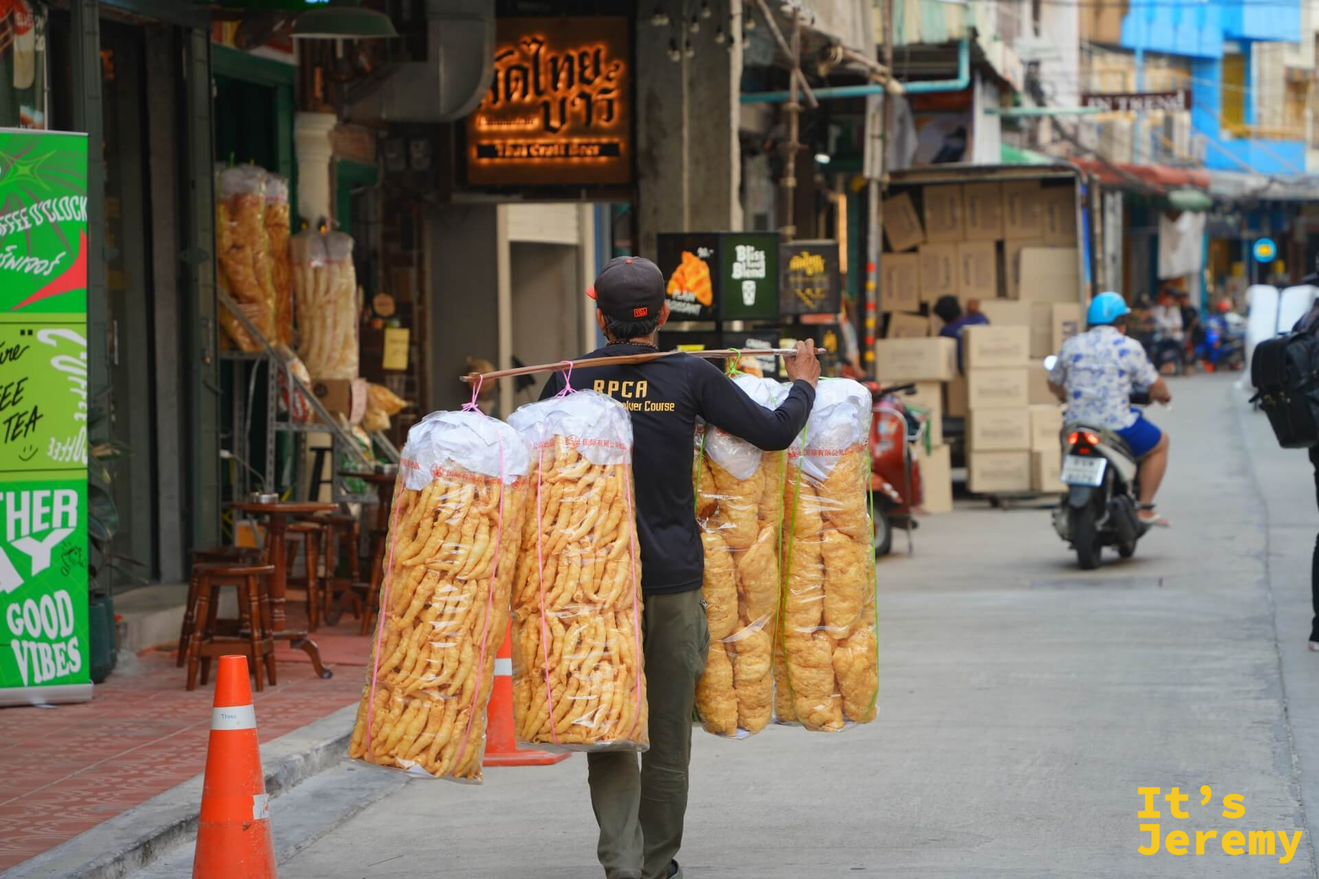 挑夫以扁擔扛著兩大袋油條行走嵩越路,背景可見「คดไทยบาร์」店舖招牌與街道日常