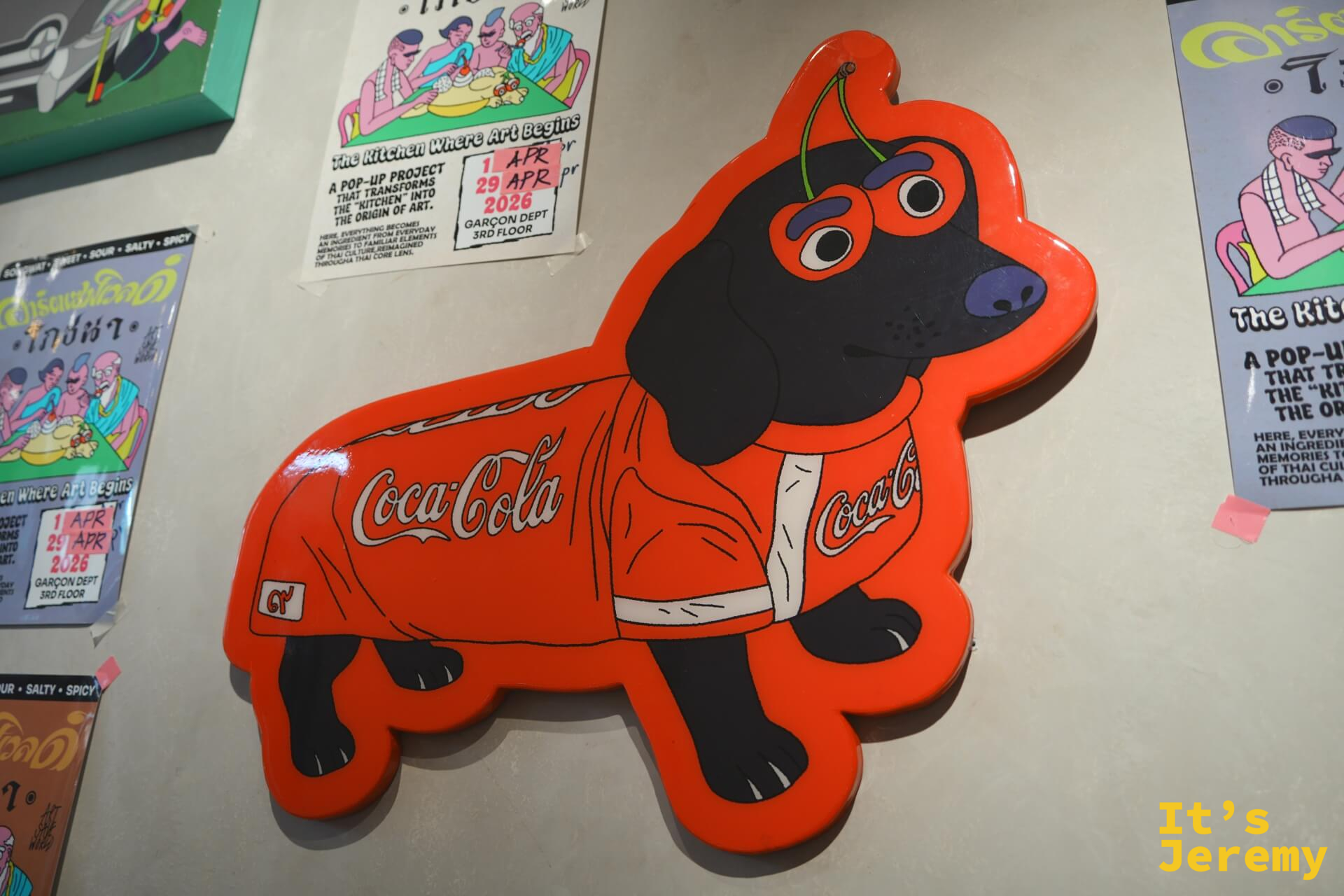 黑色臘腸犬穿著可口可樂球衣的牆面裝置藝術,旁邊貼著「The Kitchen Where Art Begins」展覽資訊