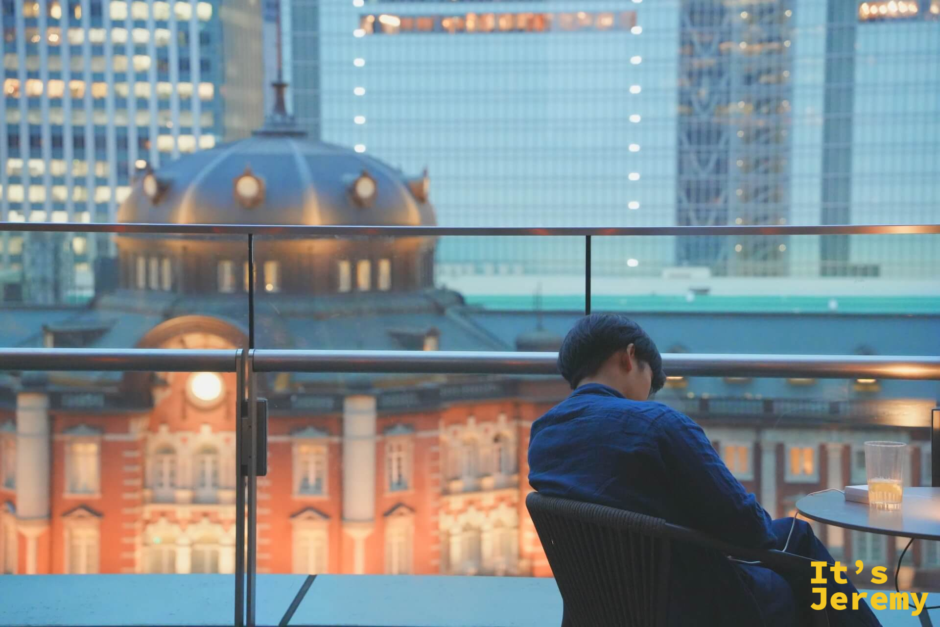 傍晚露台上,一人靜靜低頭,背景是東京車站圓頂在玻璃帷幕群中的剪影
