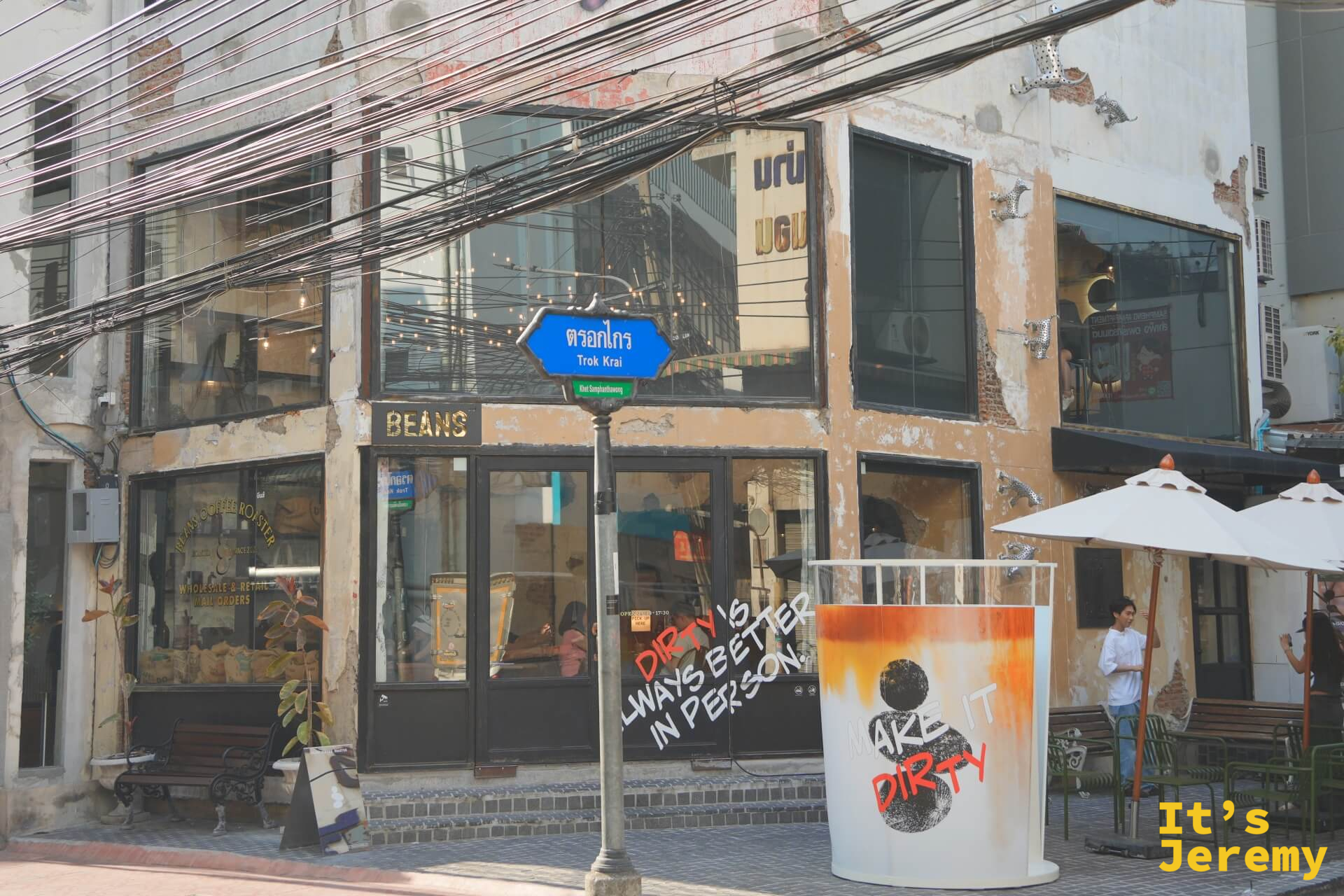 BEANS Coffee Roaster Songwat 外觀,街角藍色路牌「Trok Krai」清晰可見,外牆塗鴉「DIRTY IS ALWAYS BETTER IN PERSON」,右側白色遮陽傘
