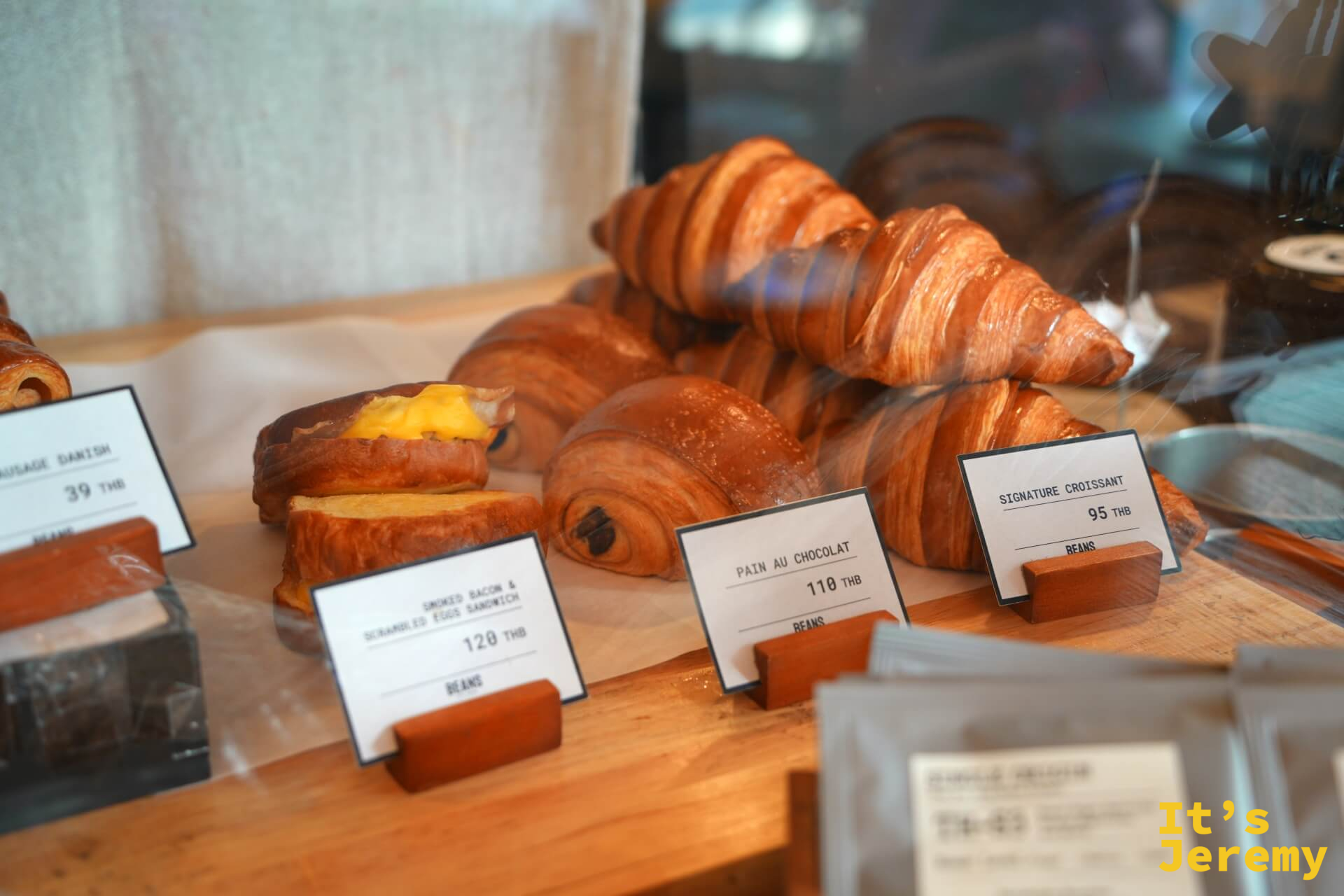玻璃展示櫃中的甜點:Signature Croissant ฿95、Pain au Chocolat ฿110、Smoked Bacon & Scrambled Eggs Sandwich ฿120、Sausage Danish ฿39