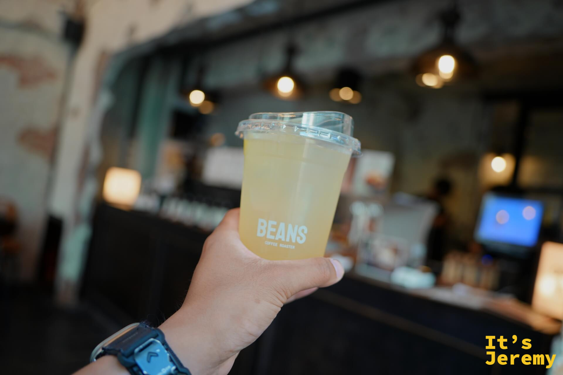 手持 BEANS 品牌透明杯,杯內為淡黃綠色柚子蘇打(Yuzu Soda),店內燈光與裸磚牆作為背景