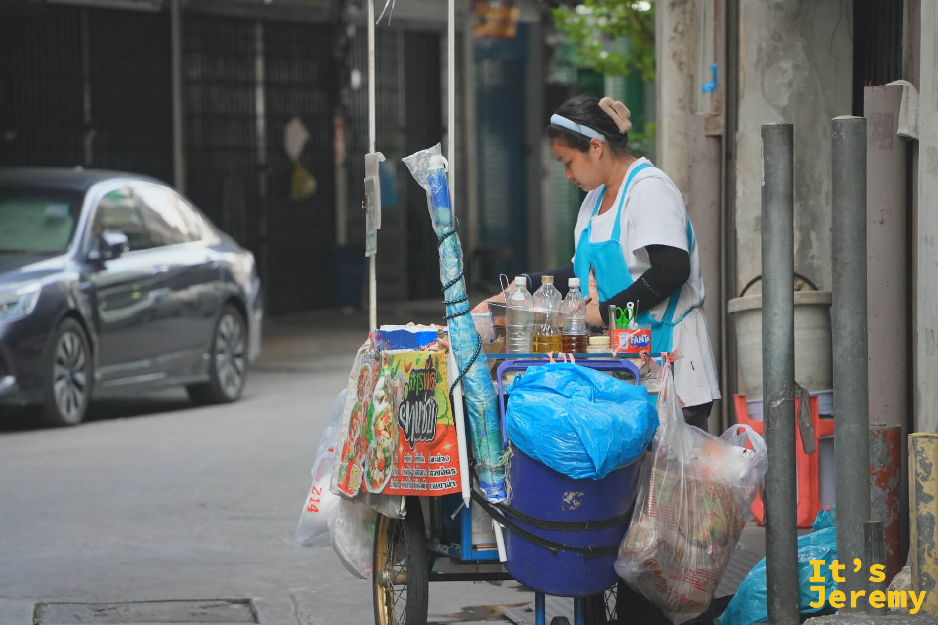 女攤主在推車旁整理食材和袋裝物品,推車上有各式飲料和調味料