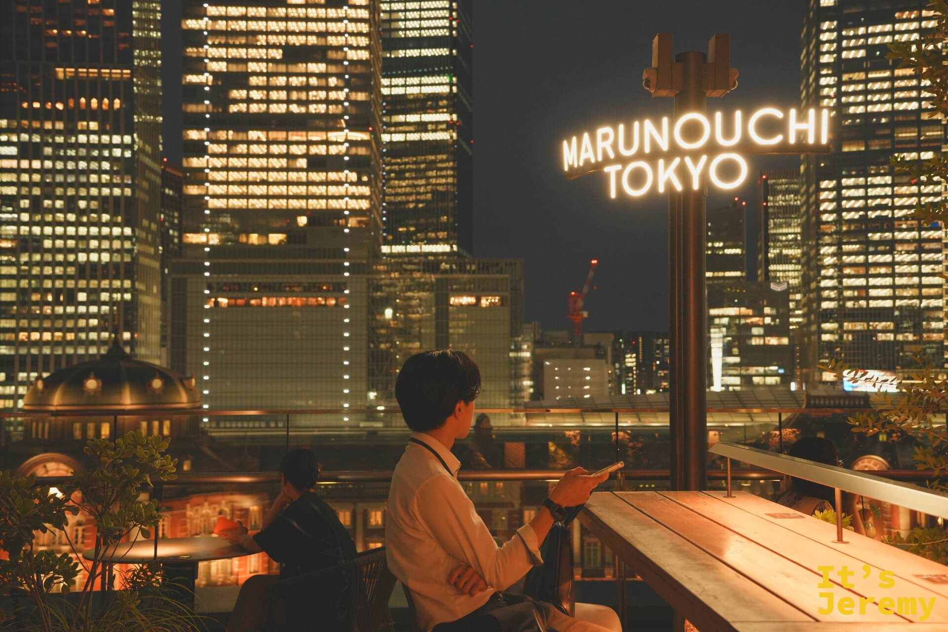 夜晚露台上,男性靠著 MARUNOUCHI TOKYO 霓虹燈牌,背後是整個丸之內的入夜景色