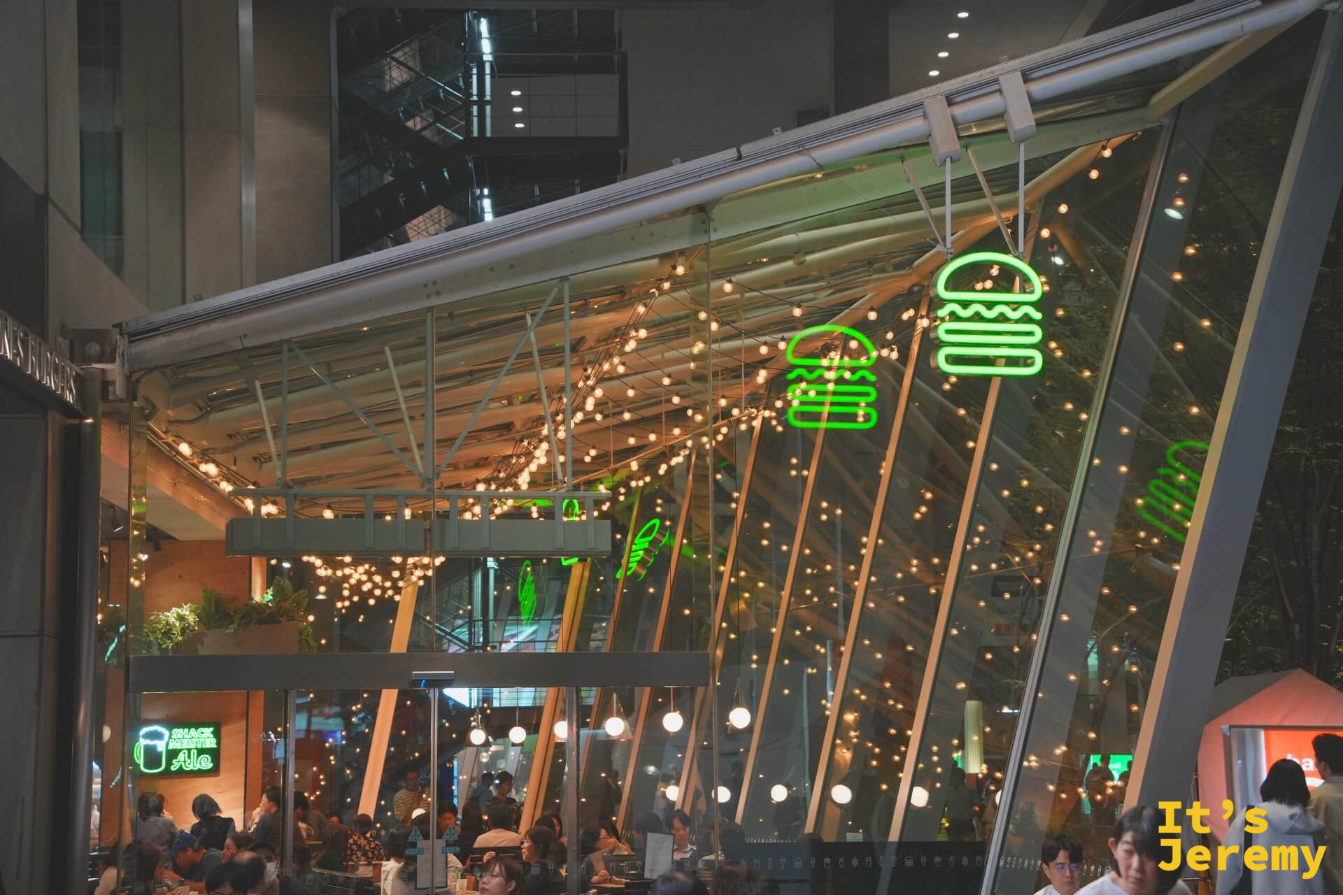 東京國際論壇廣場夜景，Shake Shack 戶外座位區在串燈與霓虹漢堡 logo 的映照下，客人陸續入座