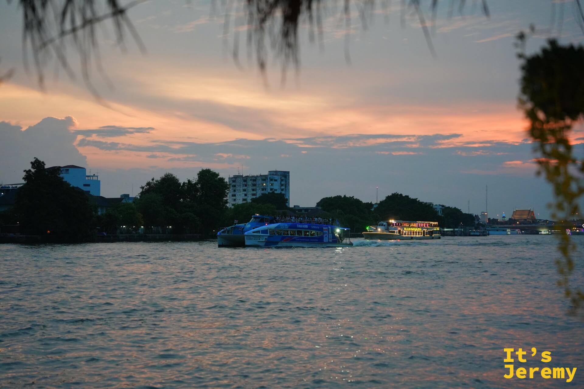 傍晚的昭披耶河，橙色夕陽映在水面，渡輪與觀光船緩緩駛過，河對岸市區燈光漸亮