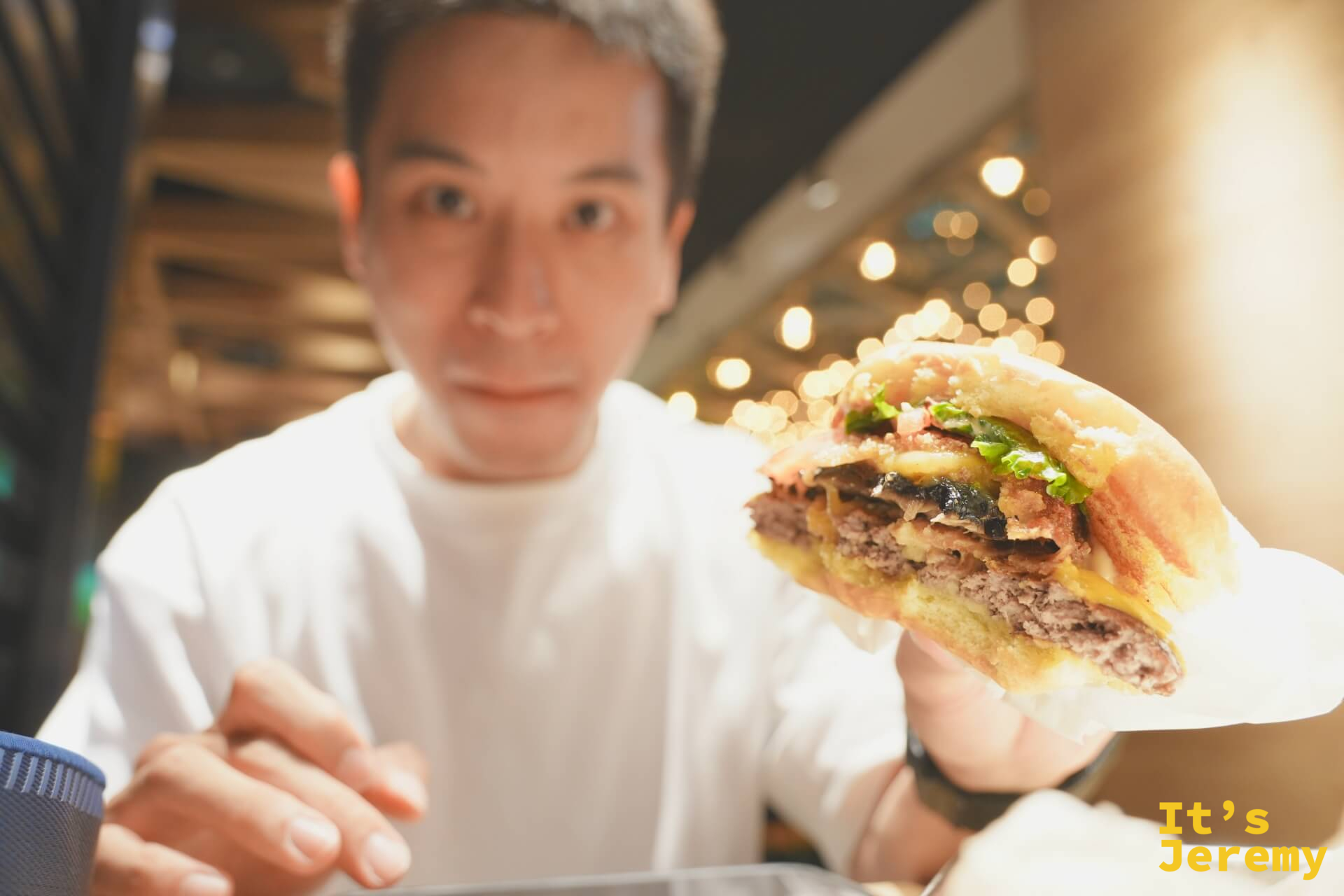 Jeremy 手持咬了一口的 ShackStack 漢堡，截面清楚顯示牛肉、起司、炸波特貝羅蘑菇的三層結構，背景是餐廳暖色燈光 bokeh