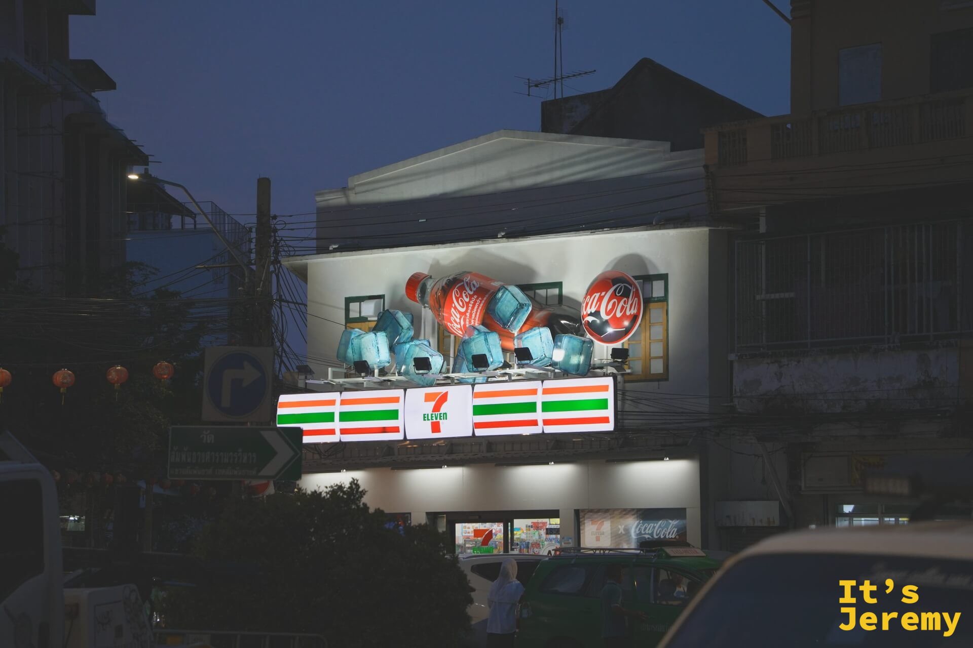 入夜後的 7-Eleven 建築:立體 Coca-Cola 玻璃瓶裝置在燈光下,左側可見紅燈籠