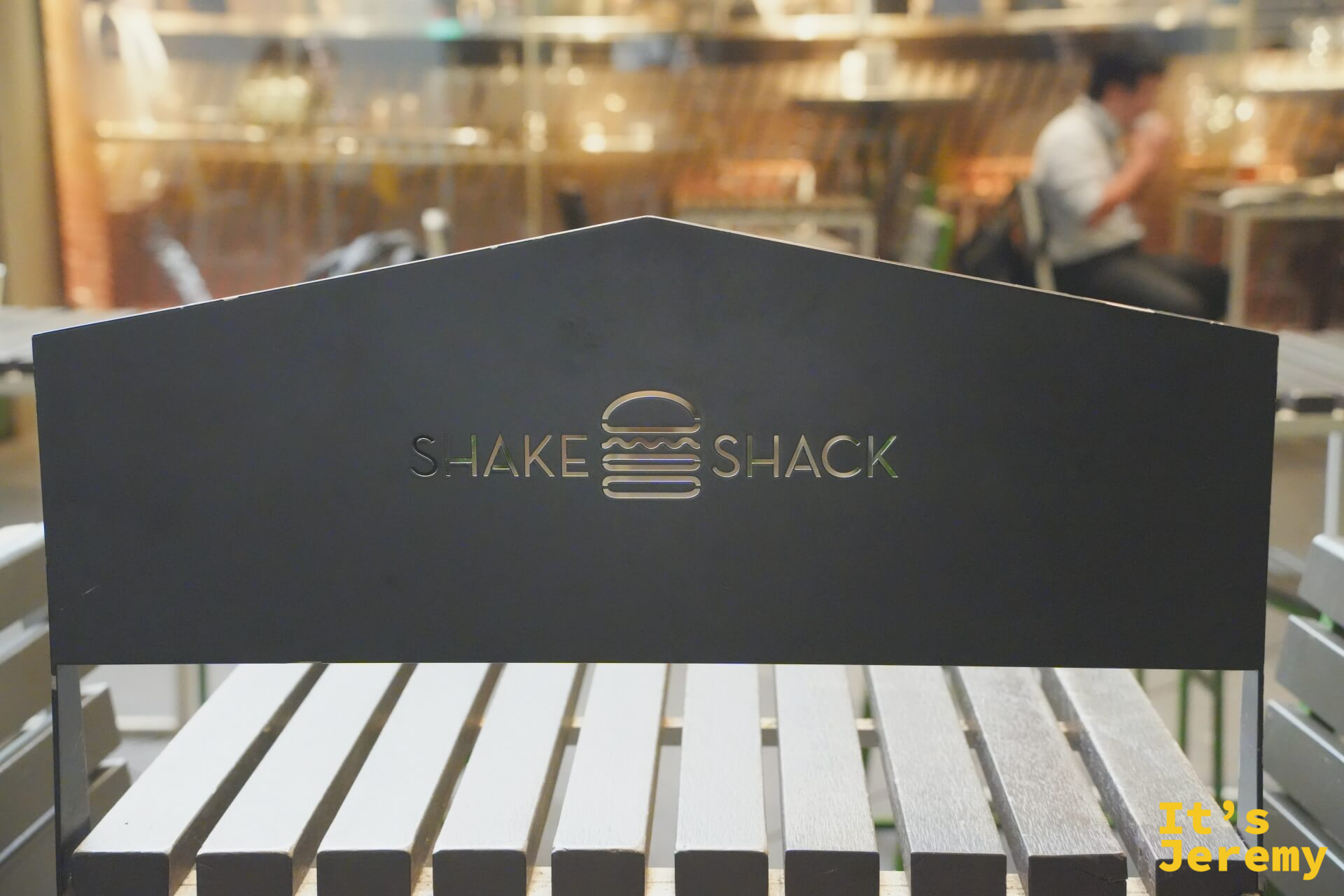 戶外廣場木質長椅椅背上的金色 Shake Shack logo，背景是餐廳室內的暖色燈光
