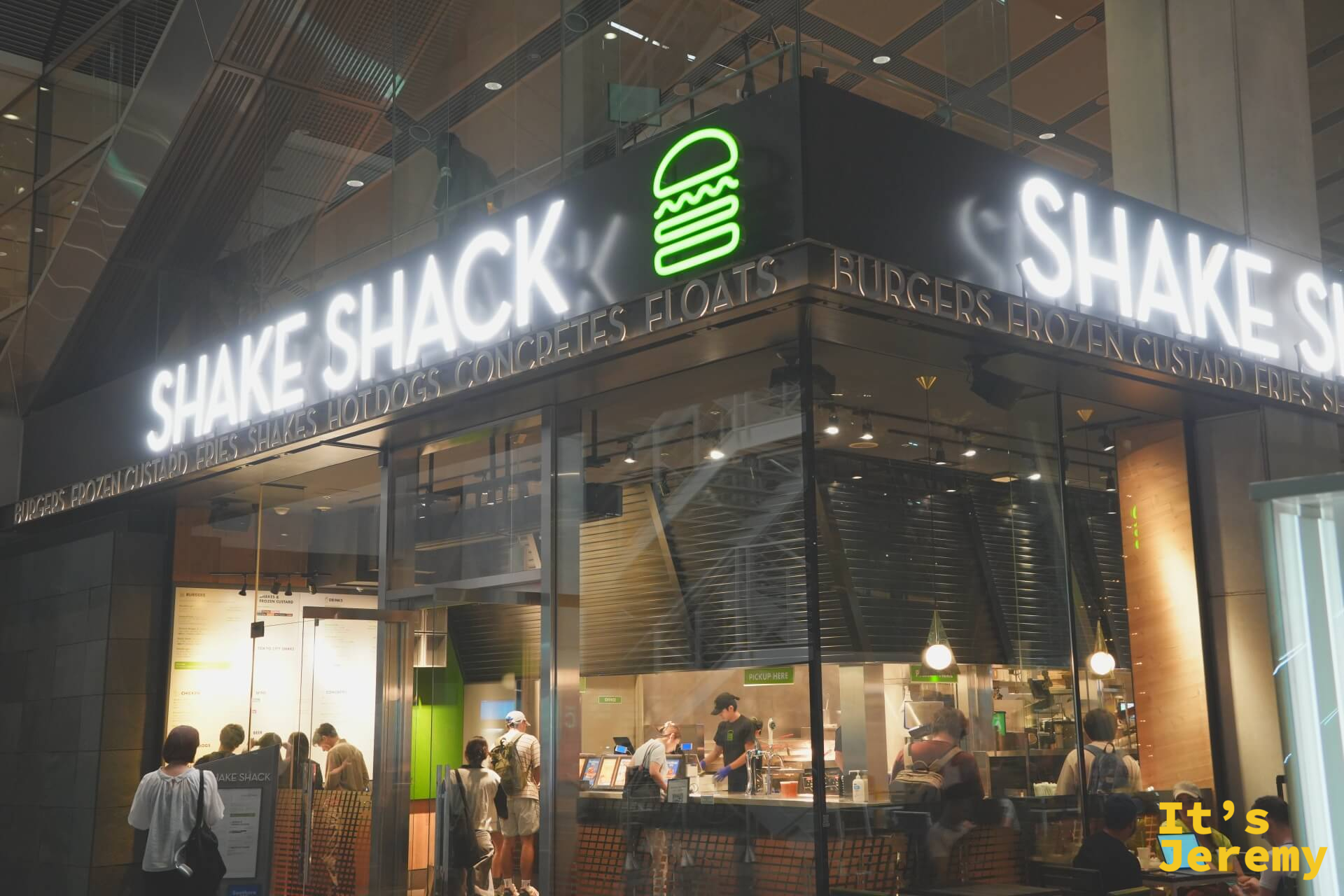 Shake Shack 東京國際論壇店夜間全景，玻璃門面、白色燈箱招牌與入場中的客人