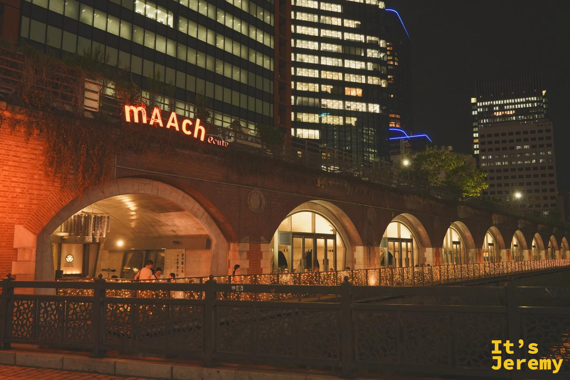mAAch ecute 神田萬世橋夜間外觀，橙紅色霓虹招牌嵌在紅磚拱廊建築頂部，川面這側的欄杆滿是串燈裝飾，後方辦公大樓燈火通明