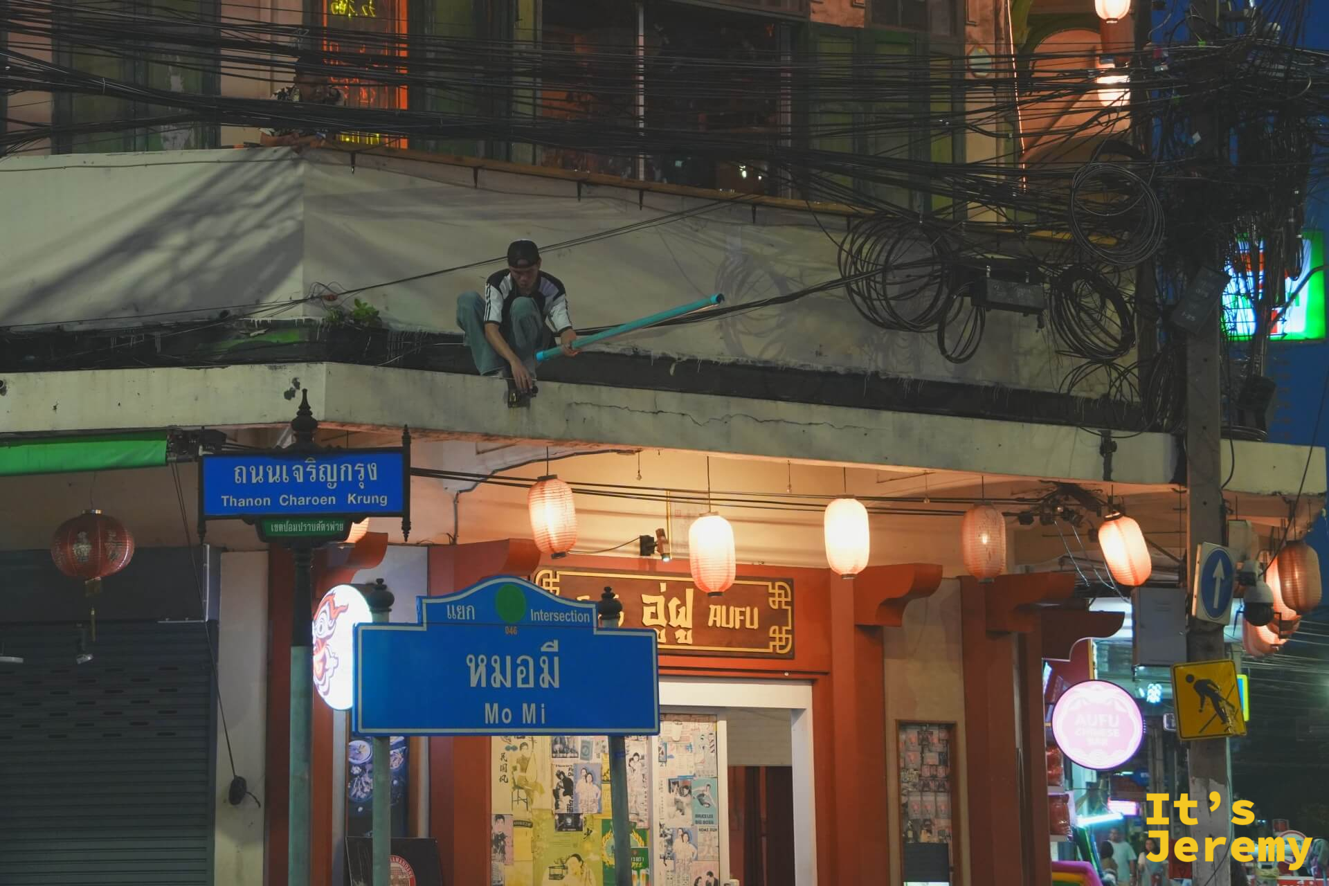 夜間路口:「Thanon Charoen Krung」路牌下方是「Mo Mi」小巷指標,一名男子趴在二樓欄杆外,附近食堂有紙燈籠