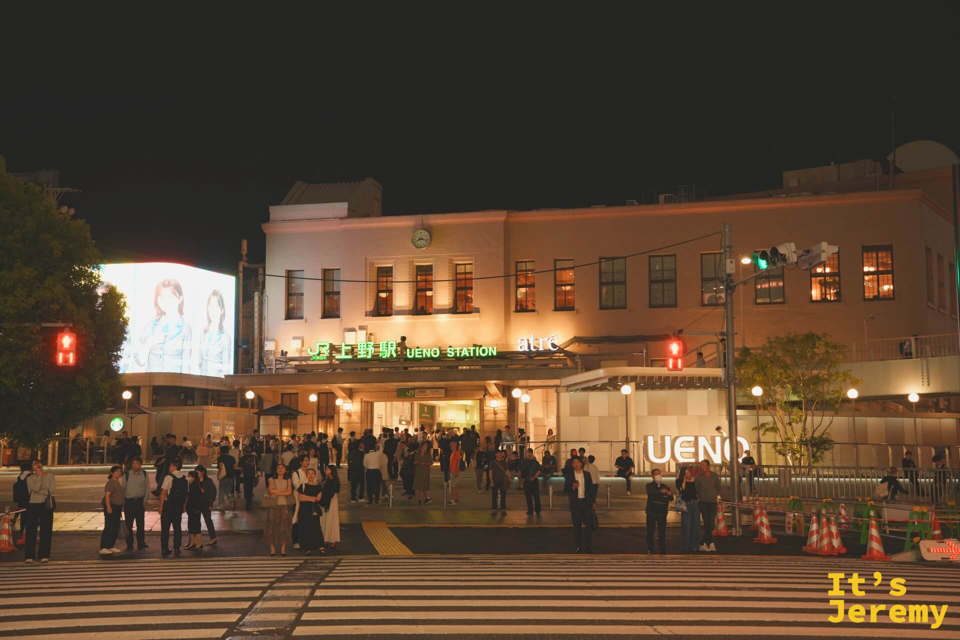 上野駅正面全景夜拍，車站白色建築打燈，UENO STATION 招牌明亮，廣場前人群聚集，廣告大螢幕在左側播放