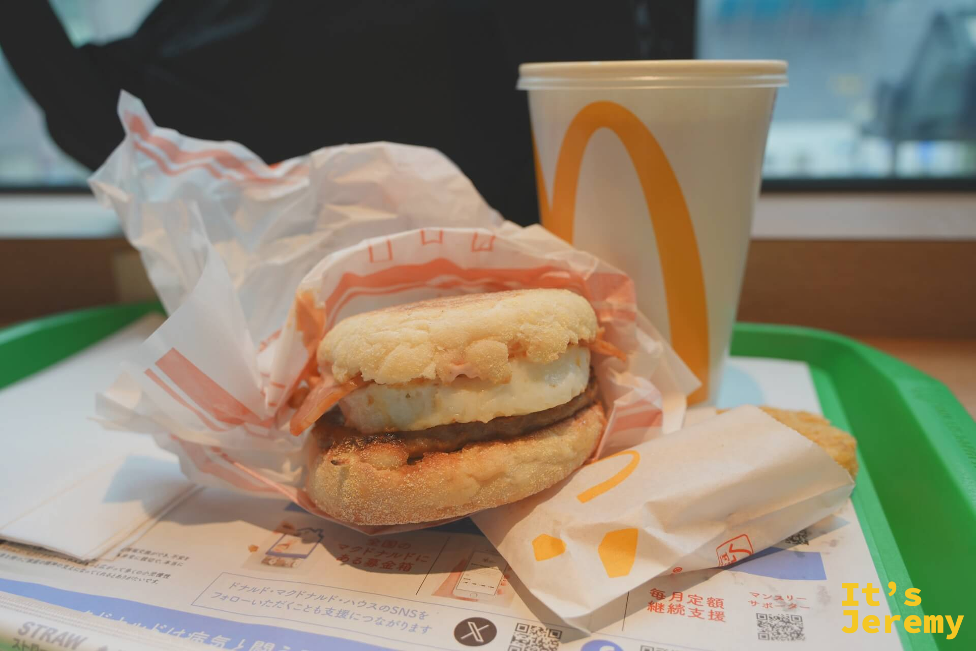 麥當勞早餐：英式鬆餅堡半開包裝放在托盤上，旁邊是紙杯飲料，底部可見日本麥當勞的宣傳單