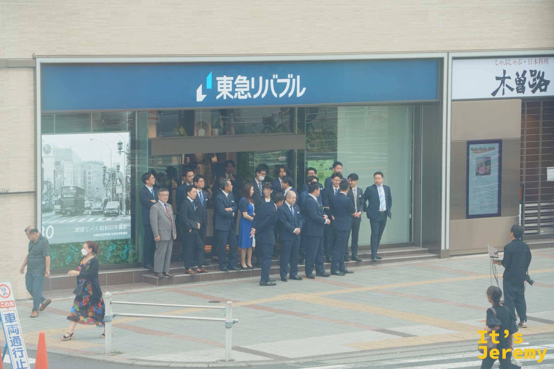 上野附近街道，一群穿西裝的商務人士在東急リバブル房仲店前排隊集合，前方有人捧著電腦充當攝影師，準備拍團體照