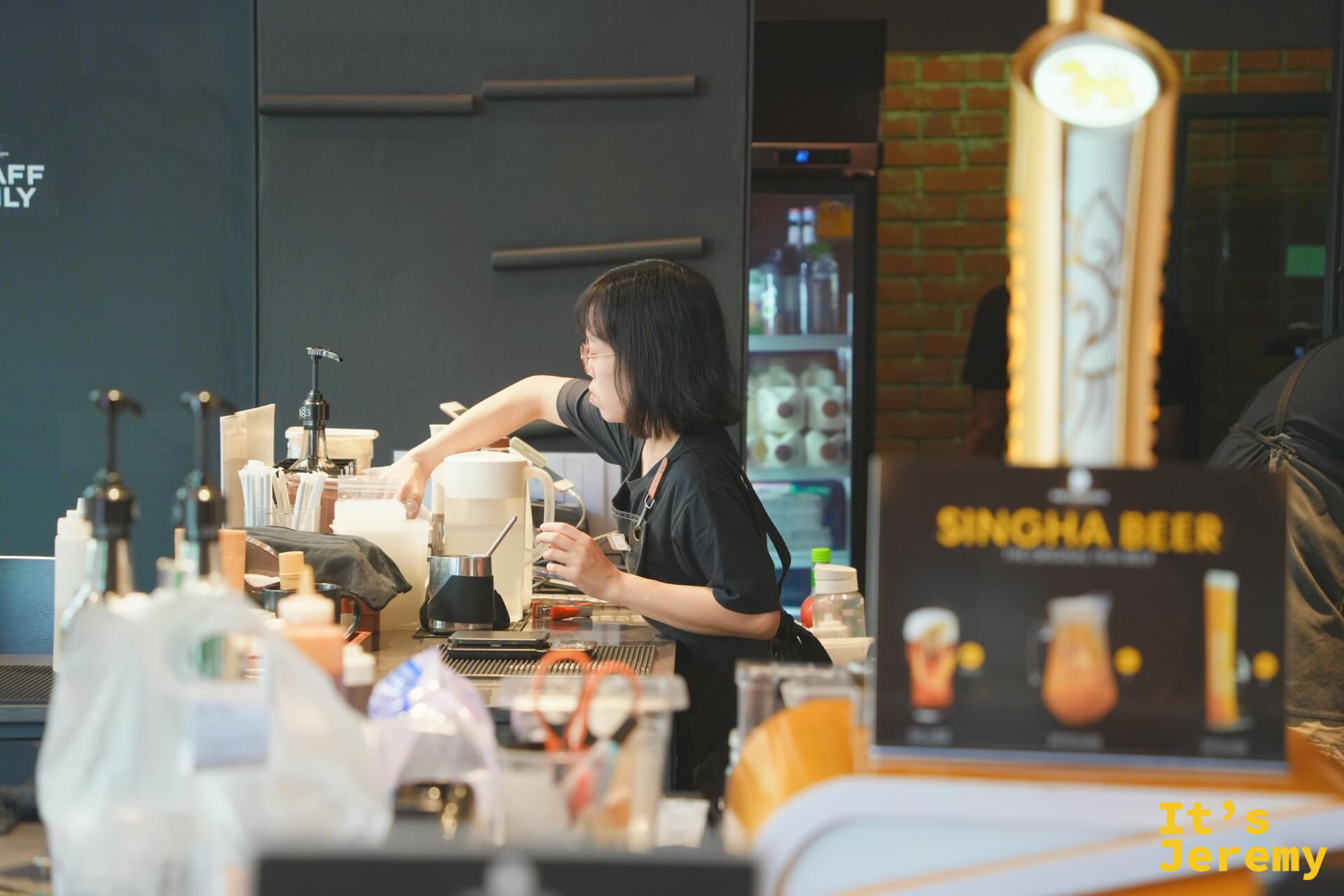 一樓女性咖啡師在吧台工作，側面鏡頭，背後有 Singha Beer 廣告立牌