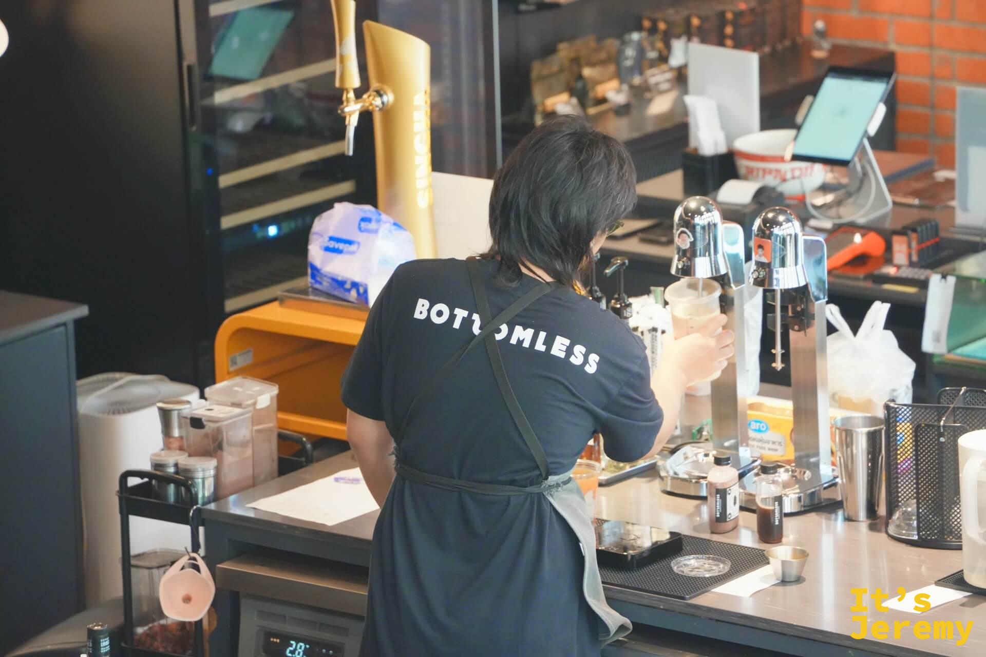 穿著 BOTTOMLESS 制服的咖啡師正在製作飲品，背面鏡頭，背後有 Singha Beer 設備