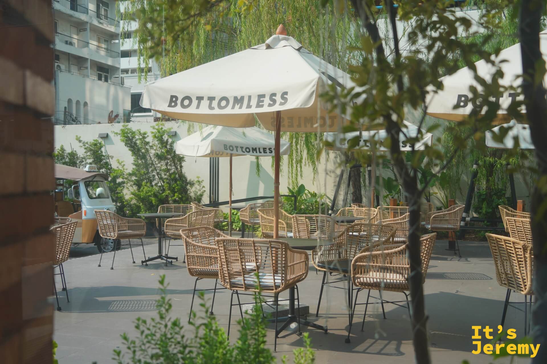 BOTTOMLESS 一樓戶外庭院，白色品牌傘、藤椅與柳樹，有柔和綠意