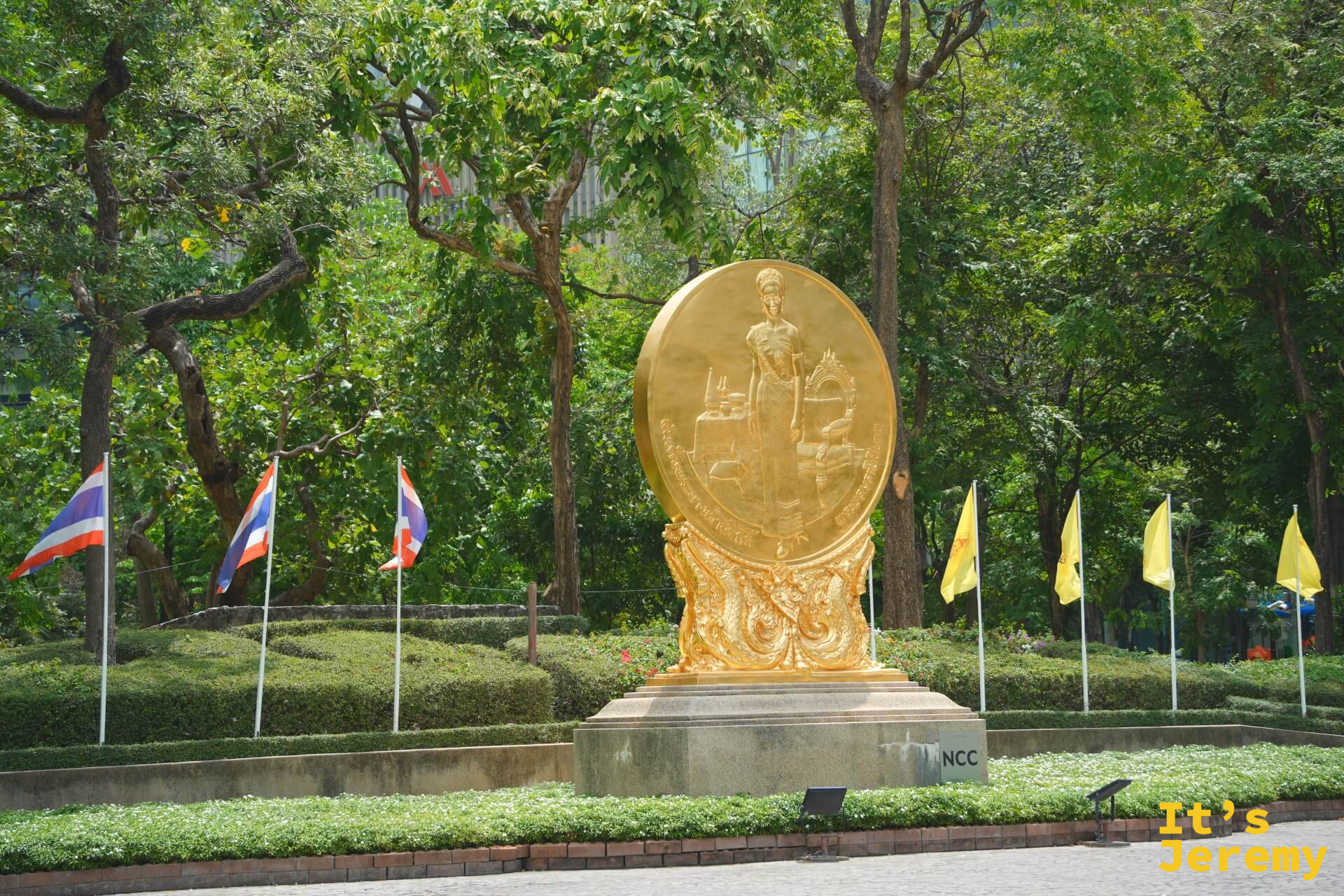 班哲希利公園入口金色圓盤浮雕紀念雕像,周圍有泰國國旗和黃旗,背景為茂密樹木